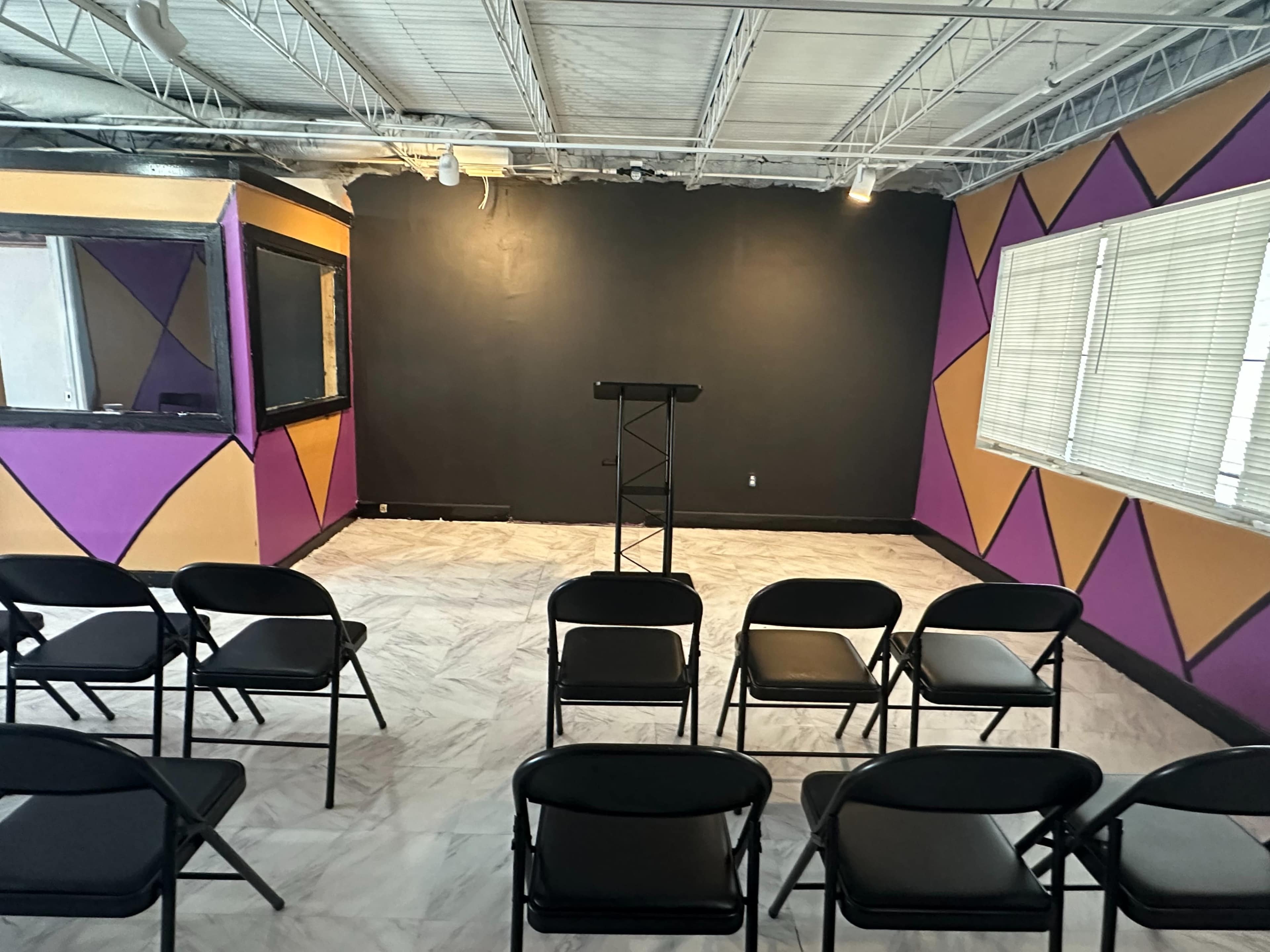 The image shows a room with black, purple, and gold geometric walls, featuring a podium at the front and several black folding chairs arranged in rows facing it.
