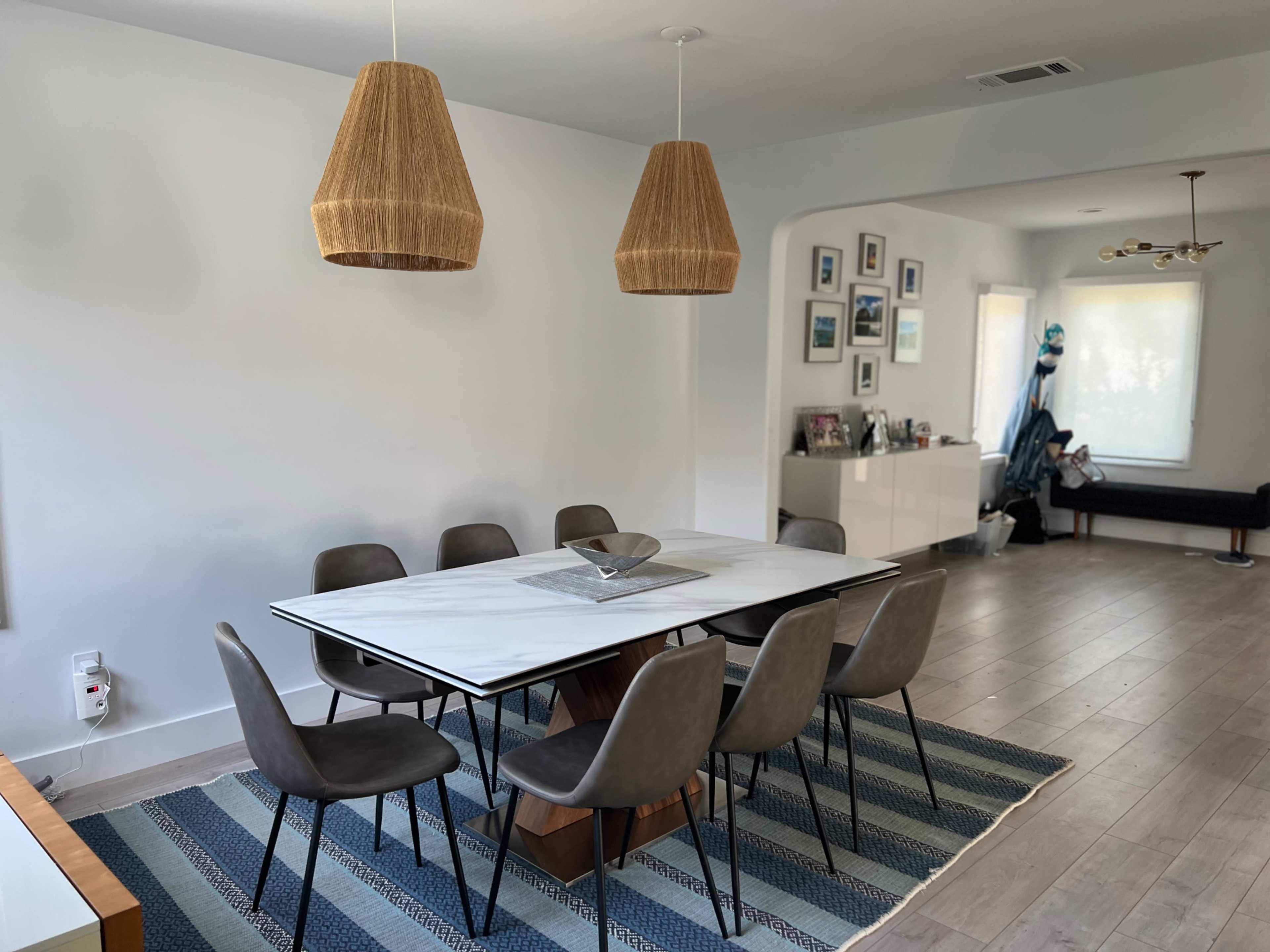A dining area features a marble-topped table surrounded by eight chairs, with two woven pendant lights hanging above and a rug underneath.