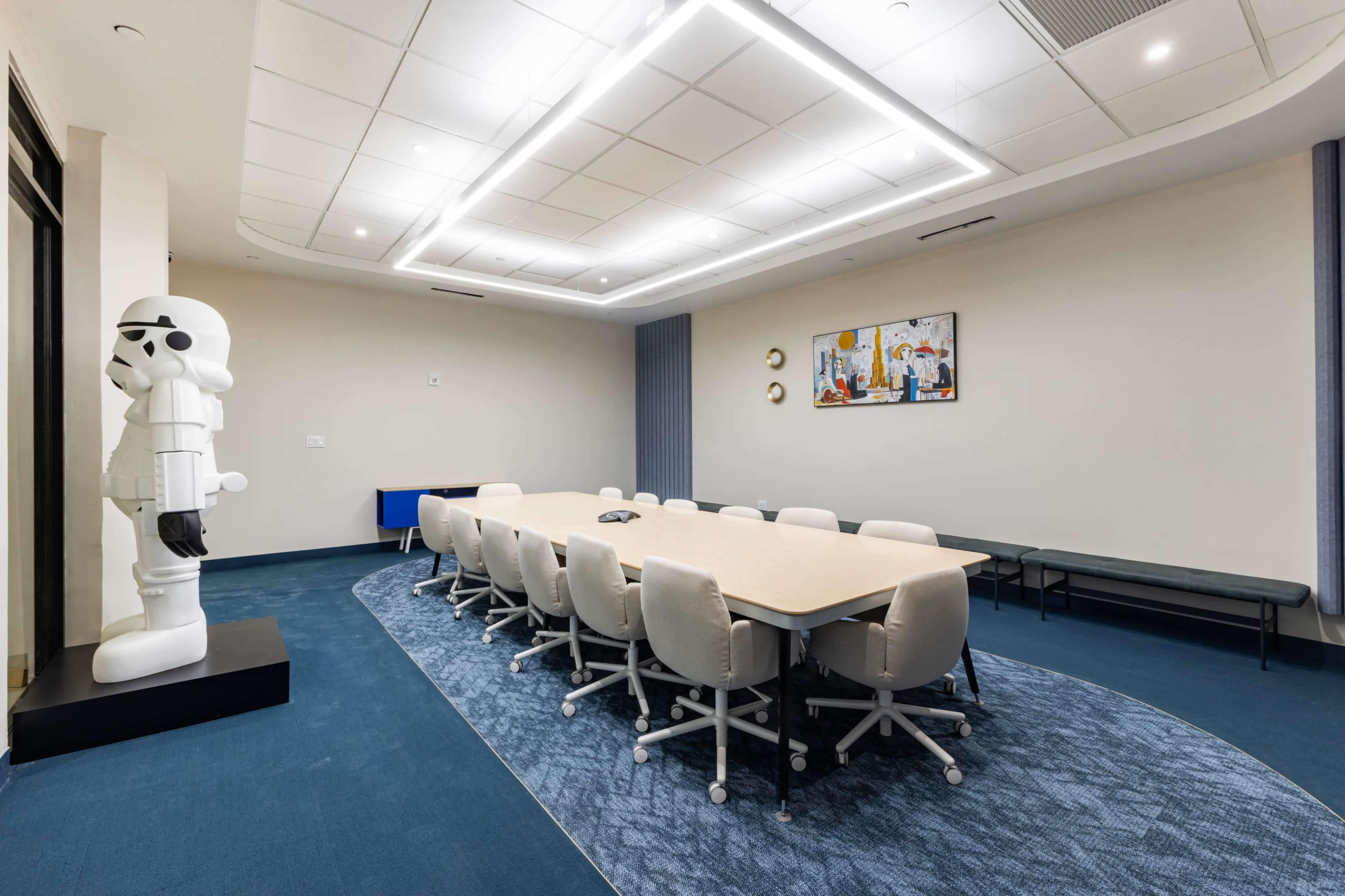 A modern conference room features a long table surrounded by upholstered chairs, with a decorative stormtrooper statue standing next to the entrance.