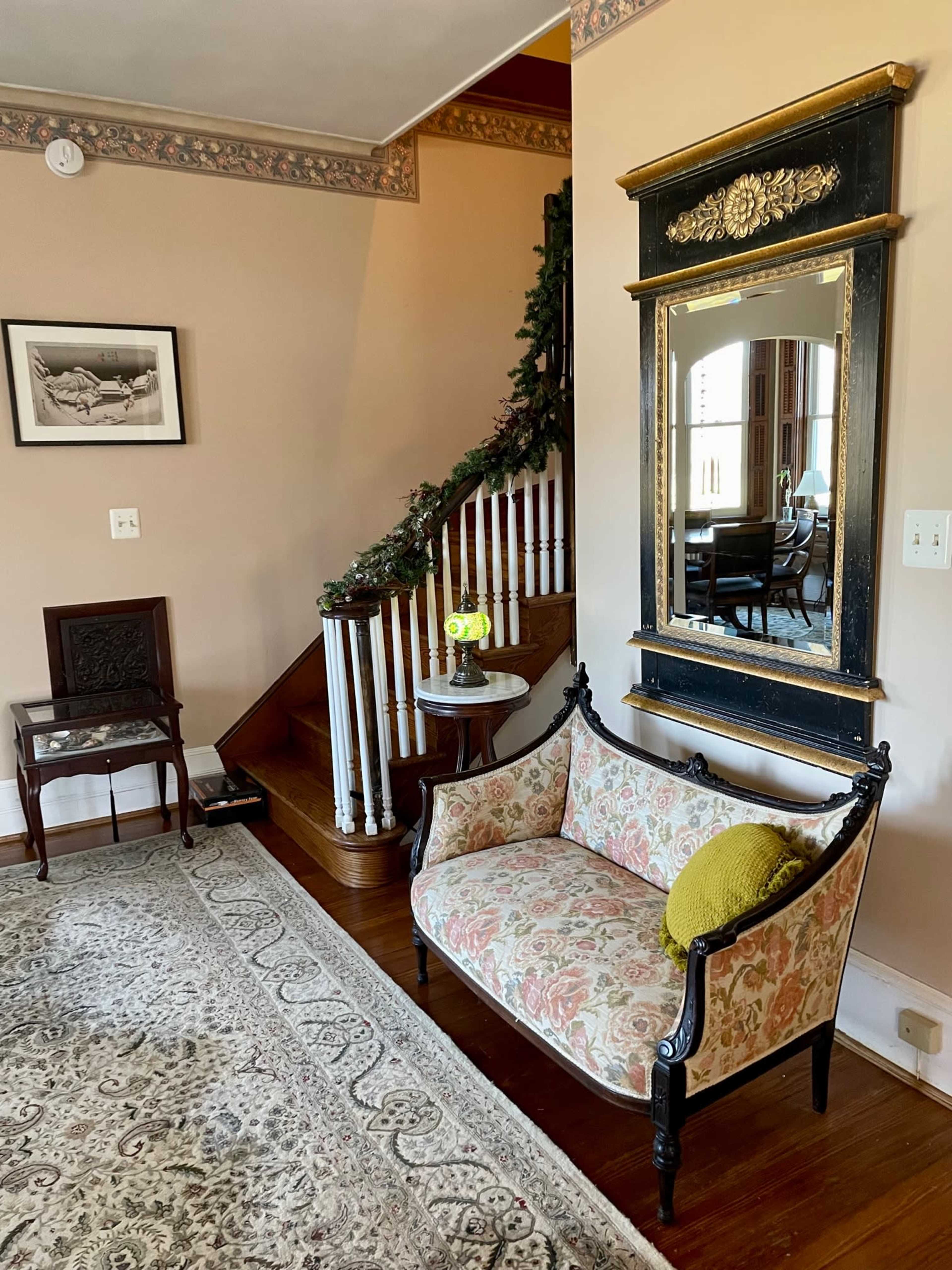 A living room area with a floral-patterned couch, a small table with a vase of flowers, a decorative mirror, and a staircase in the background.