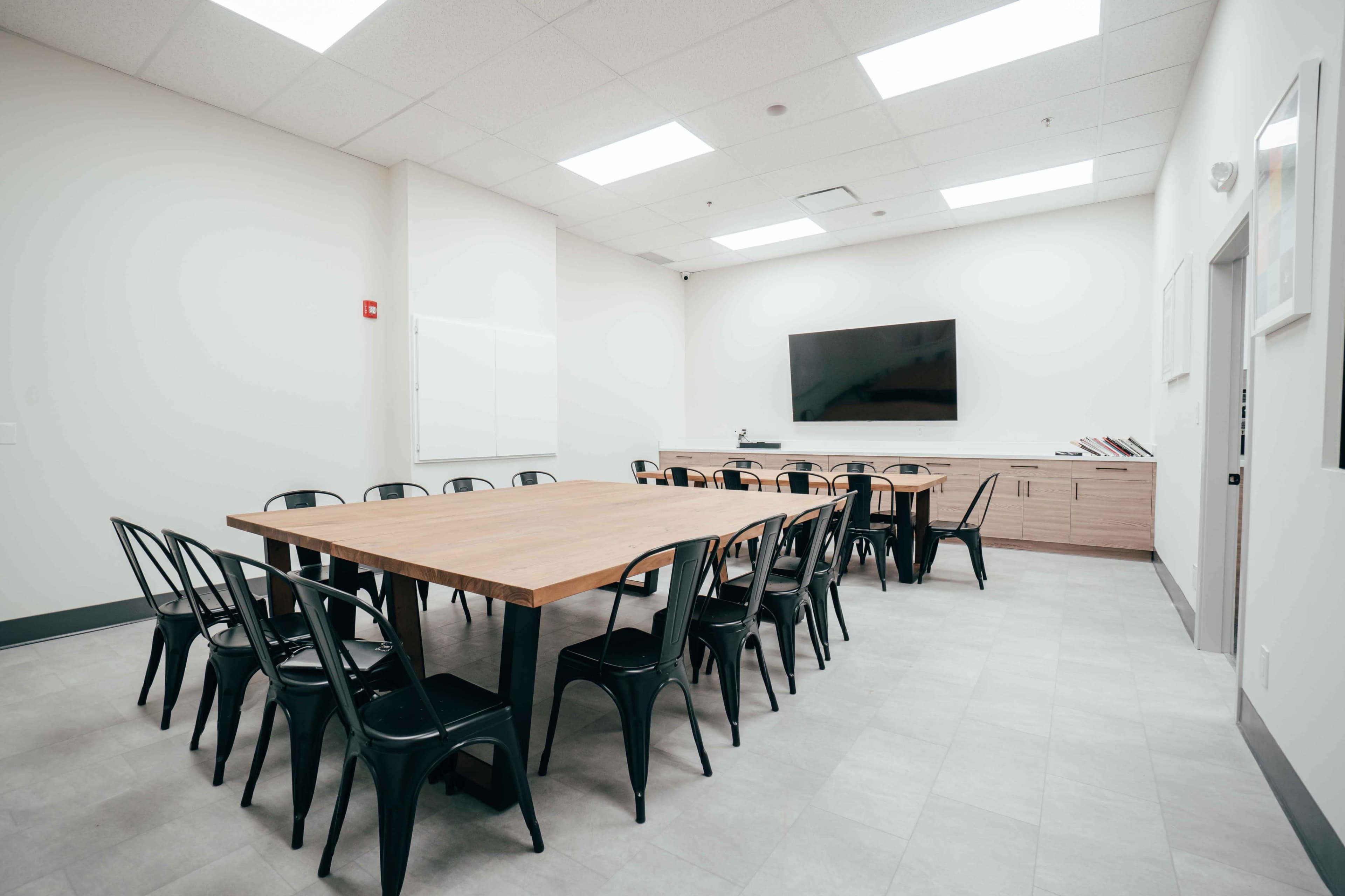 A vacant meeting room features a large wooden table surrounded by black chairs, with a wall-mounted screen and minimal furnishings.