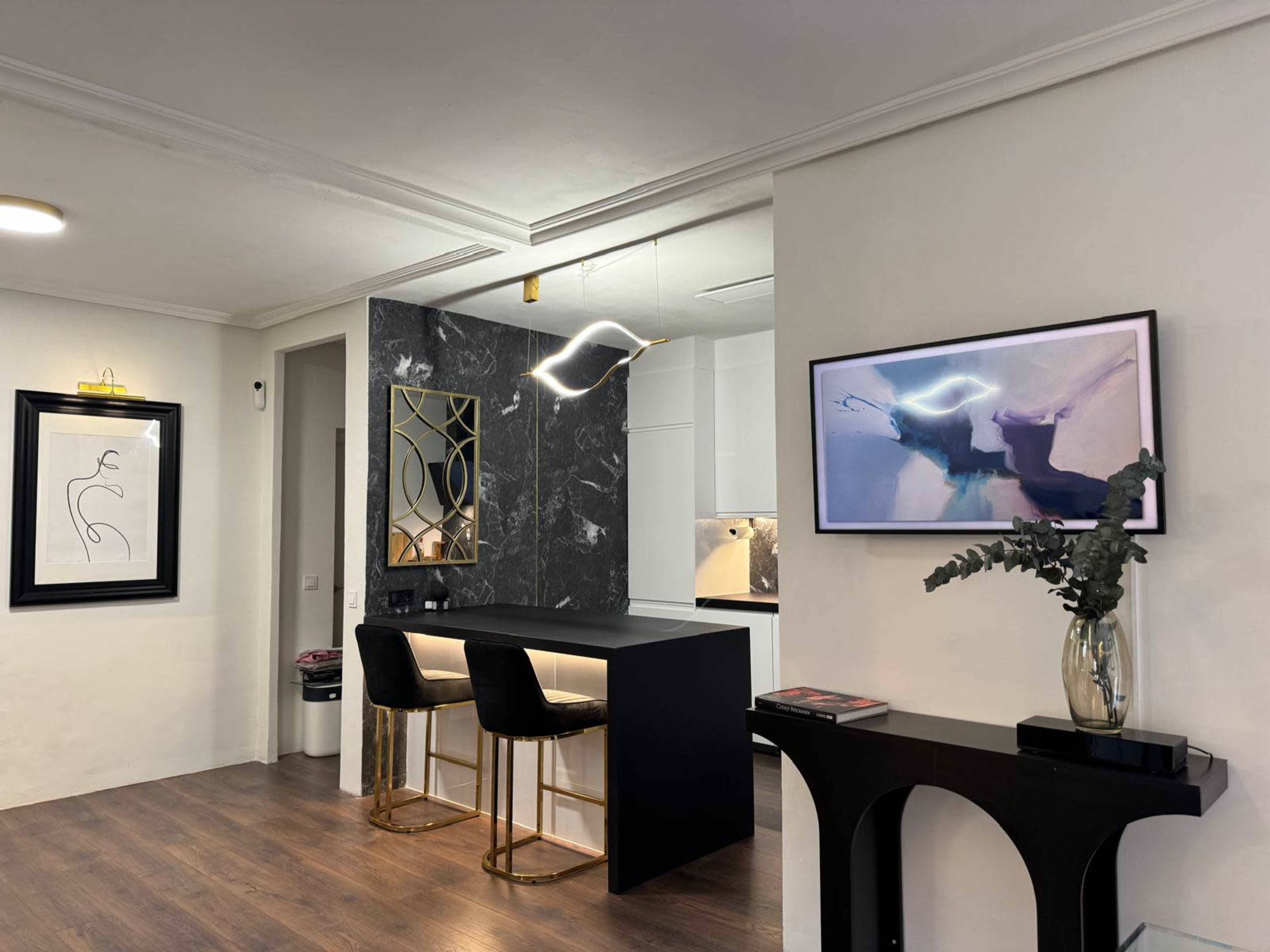 The image shows a modern kitchen area with a black island, two bar stools, a marble accent wall, and contemporary artwork framed on the walls.