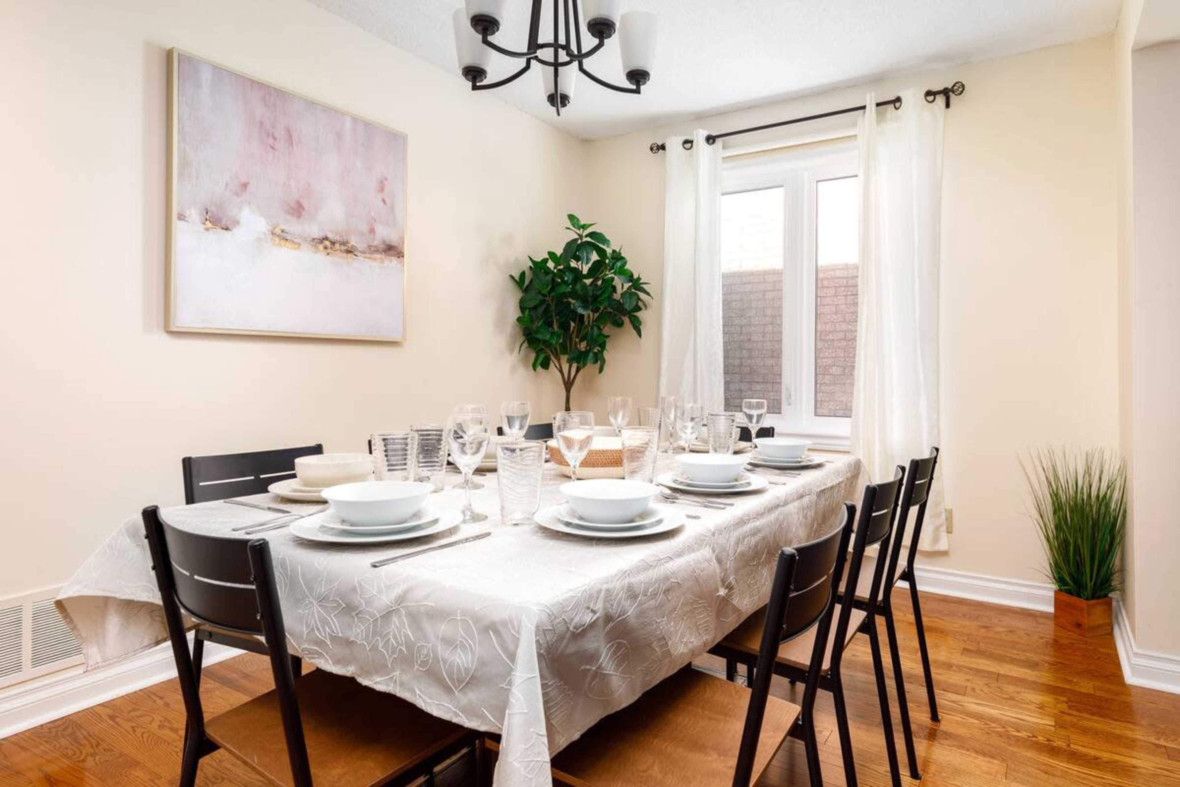 A dining table is set for a meal, featuring plates, glasses, and cutlery, illuminated by a chandelier in a light-colored room.