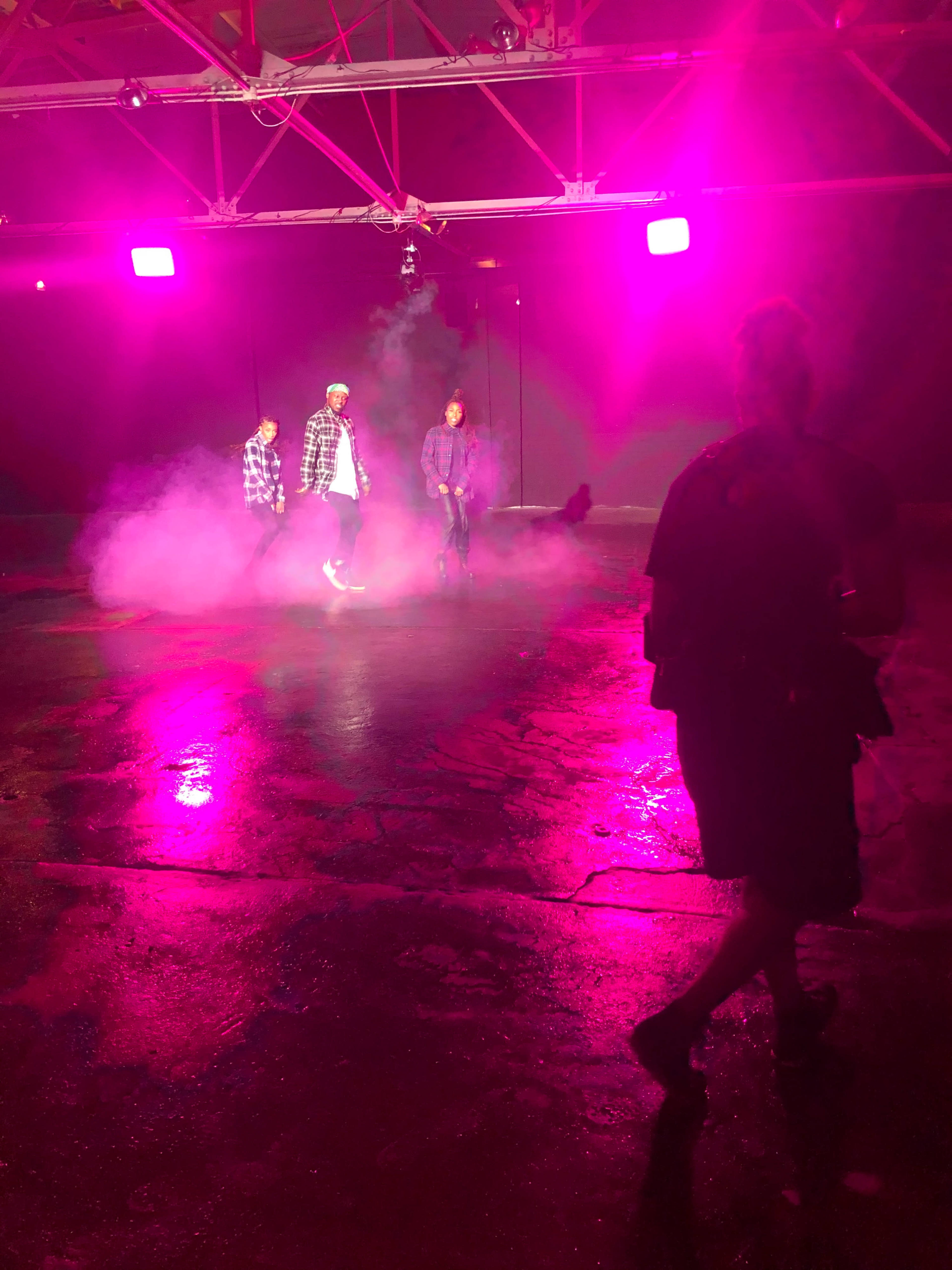 Three individuals stand on a reflective surface surrounded by pink lighting and fog.