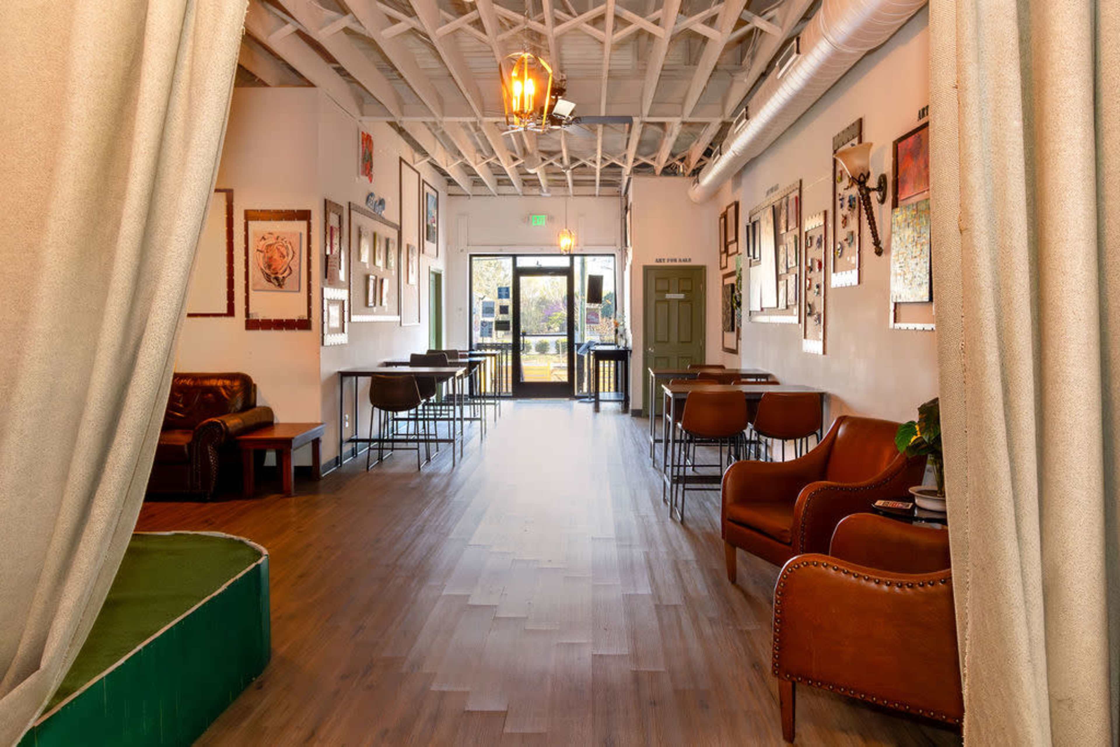The image shows a narrow interior of a café or small venue with seating areas on both sides, framed artwork on the walls, and an entrance that leads to an outdoor area.