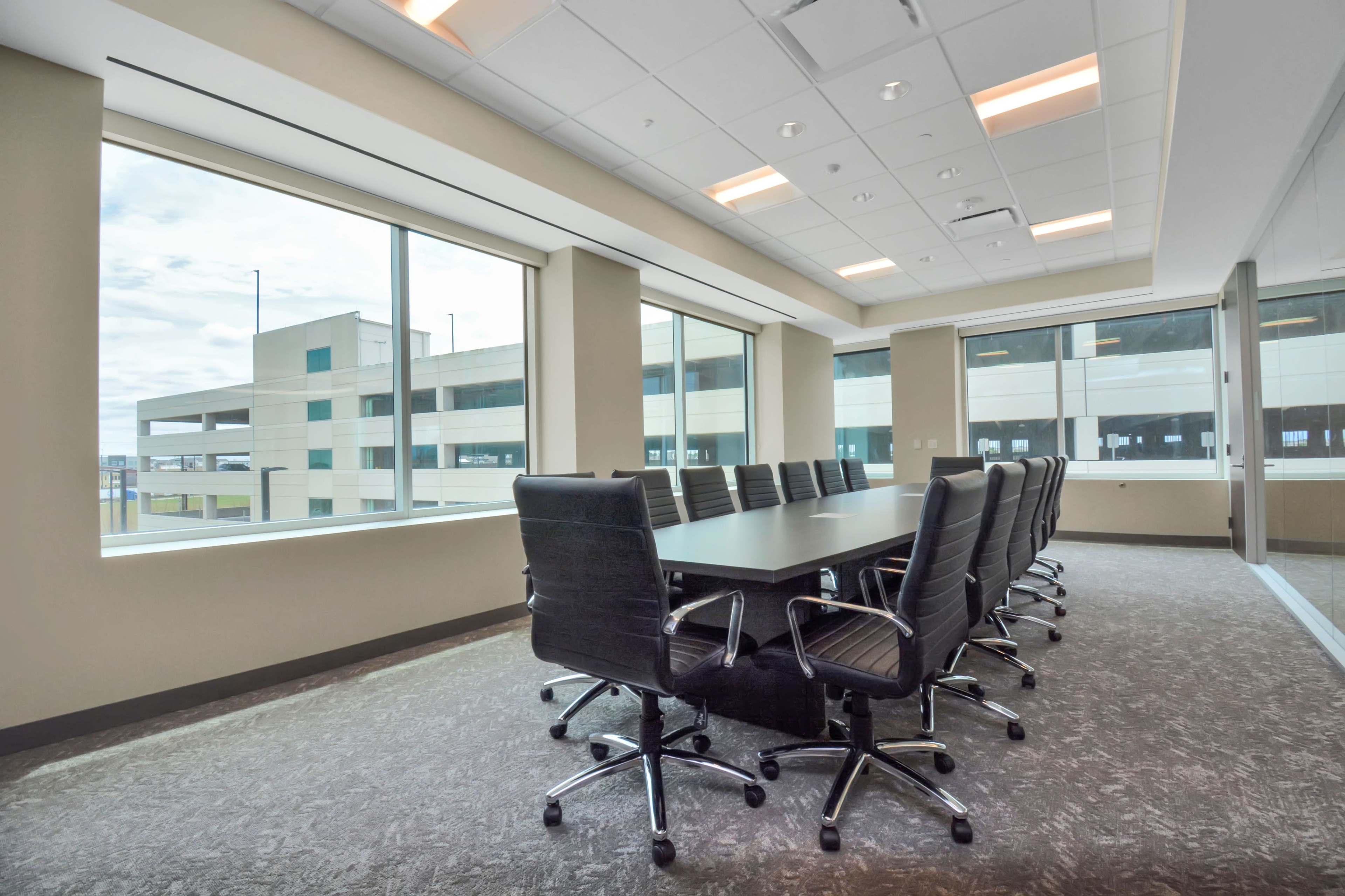 A modern conference room features a large table surrounded by black swivel chairs, illuminated by natural light from large windows.