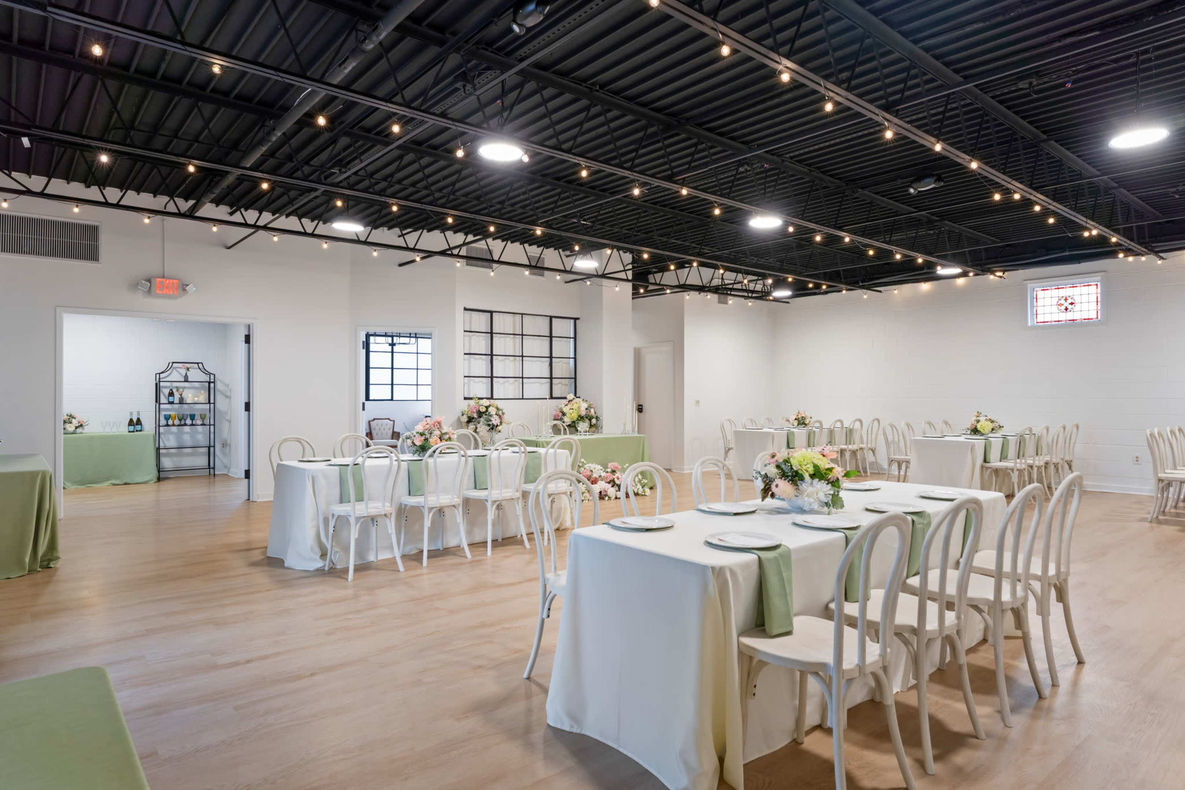 The image shows an elegantly arranged event space with white tables covered in light green runners, decorated with floral centerpieces, and string lights hanging from the ceiling.