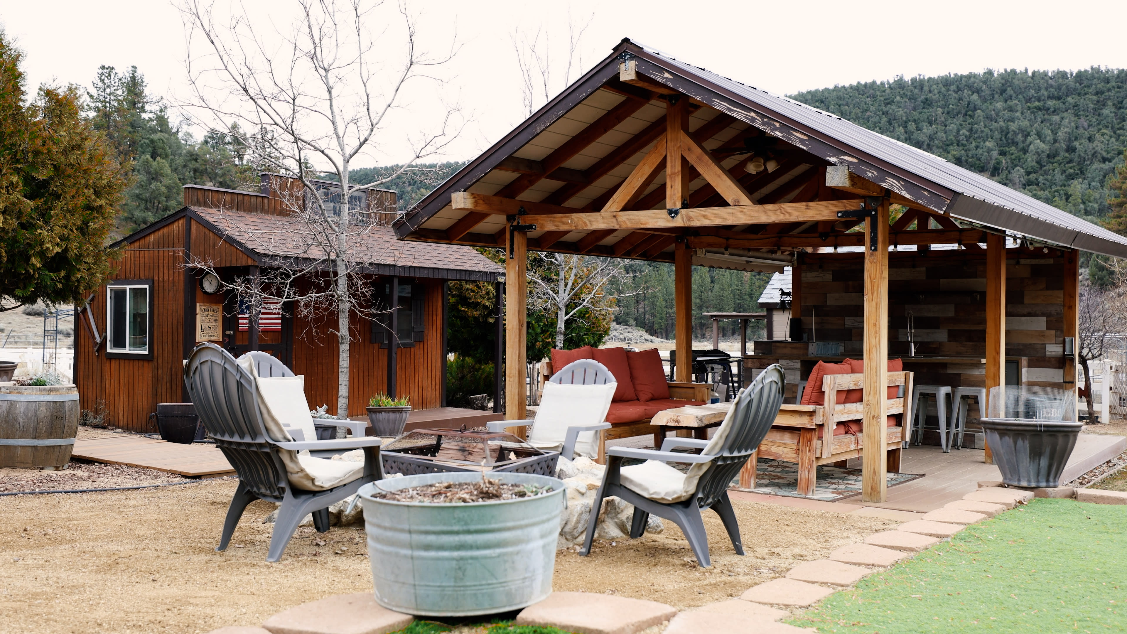 The image shows a rustic outdoor sitting area with wooden furniture and a covered pavilion, surrounded by a gravel and lawn landscape.