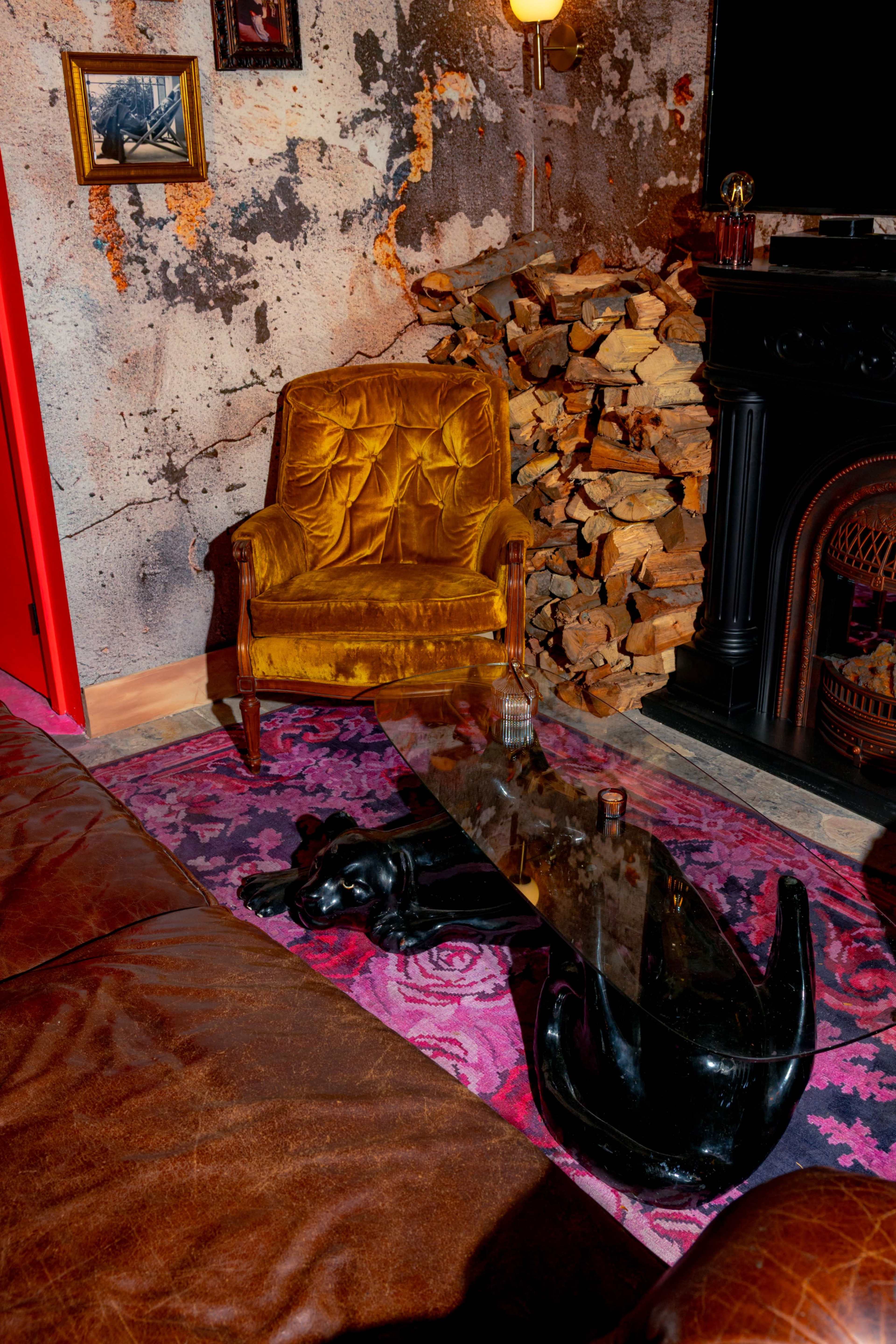 The image shows a vintage velvet armchair beside a glass coffee table supported by a black panther sculpture, set against a backdrop of textured walls and a stack of firewood.