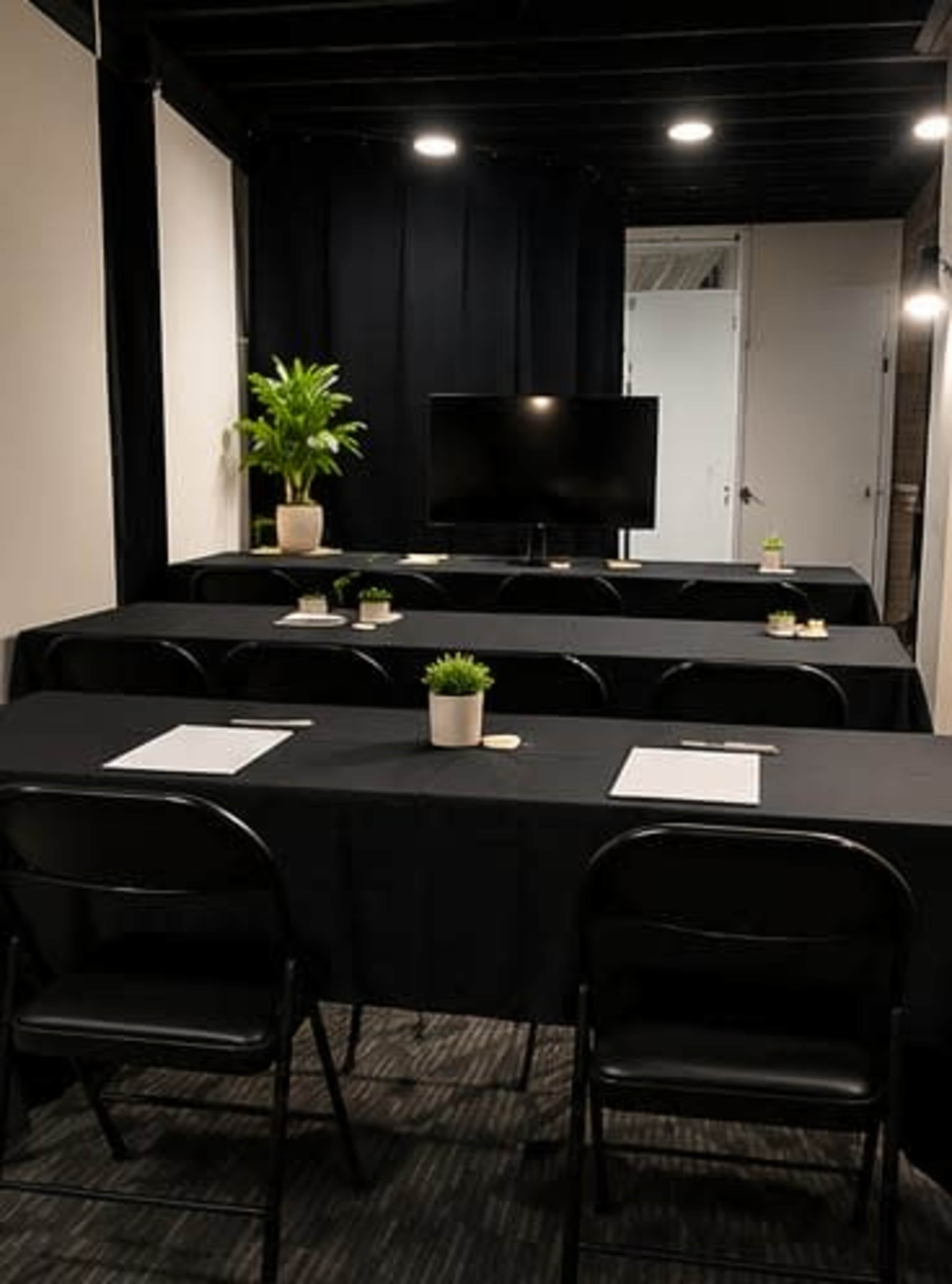 A small conference room features several black tables arranged in rows, each with a potted plant and notepads, and a television mounted on the wall.