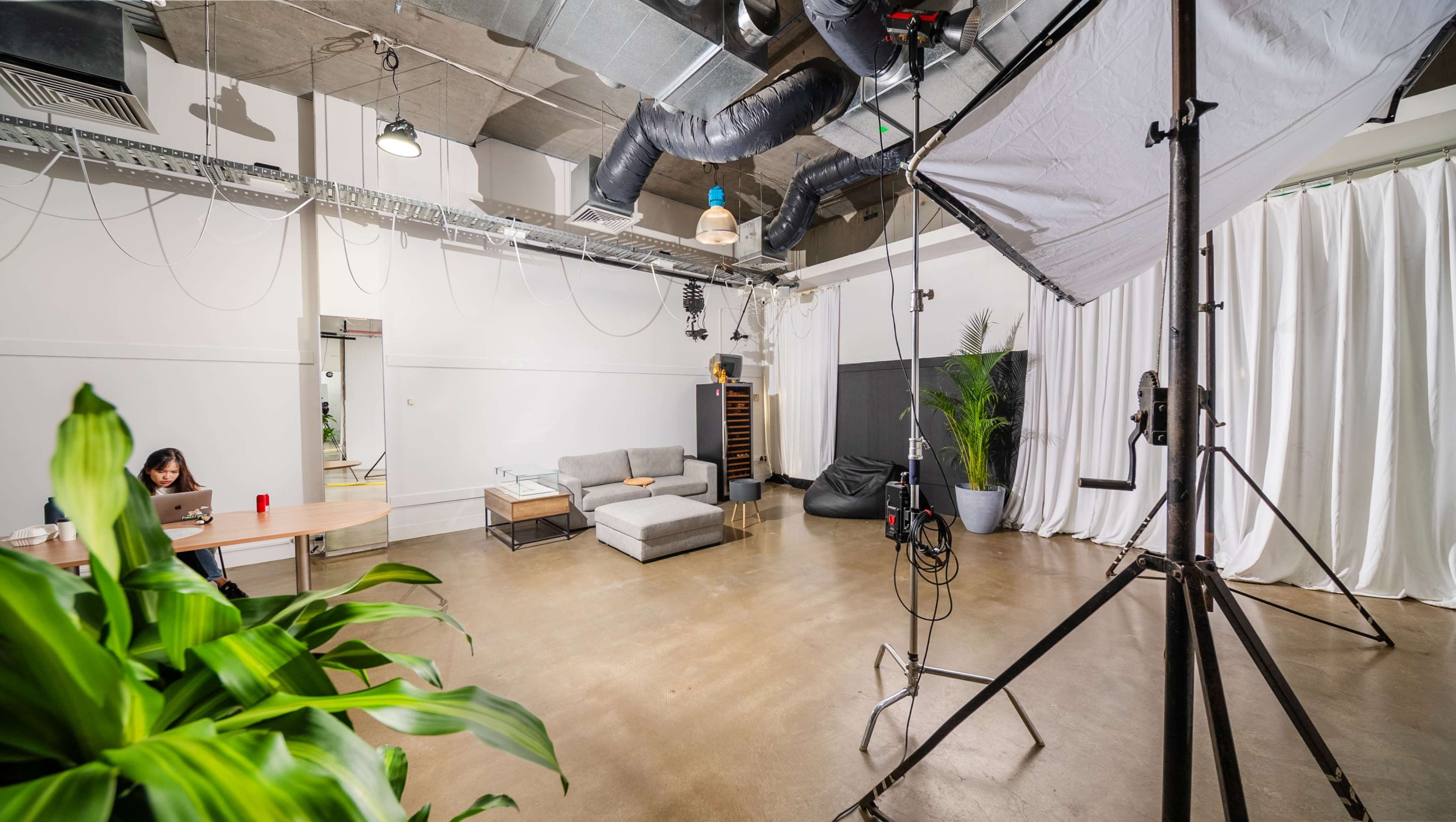 A minimalistic studio space features a couch, a table with a person working on a laptop, and a backdrop setup with a large light fixture.