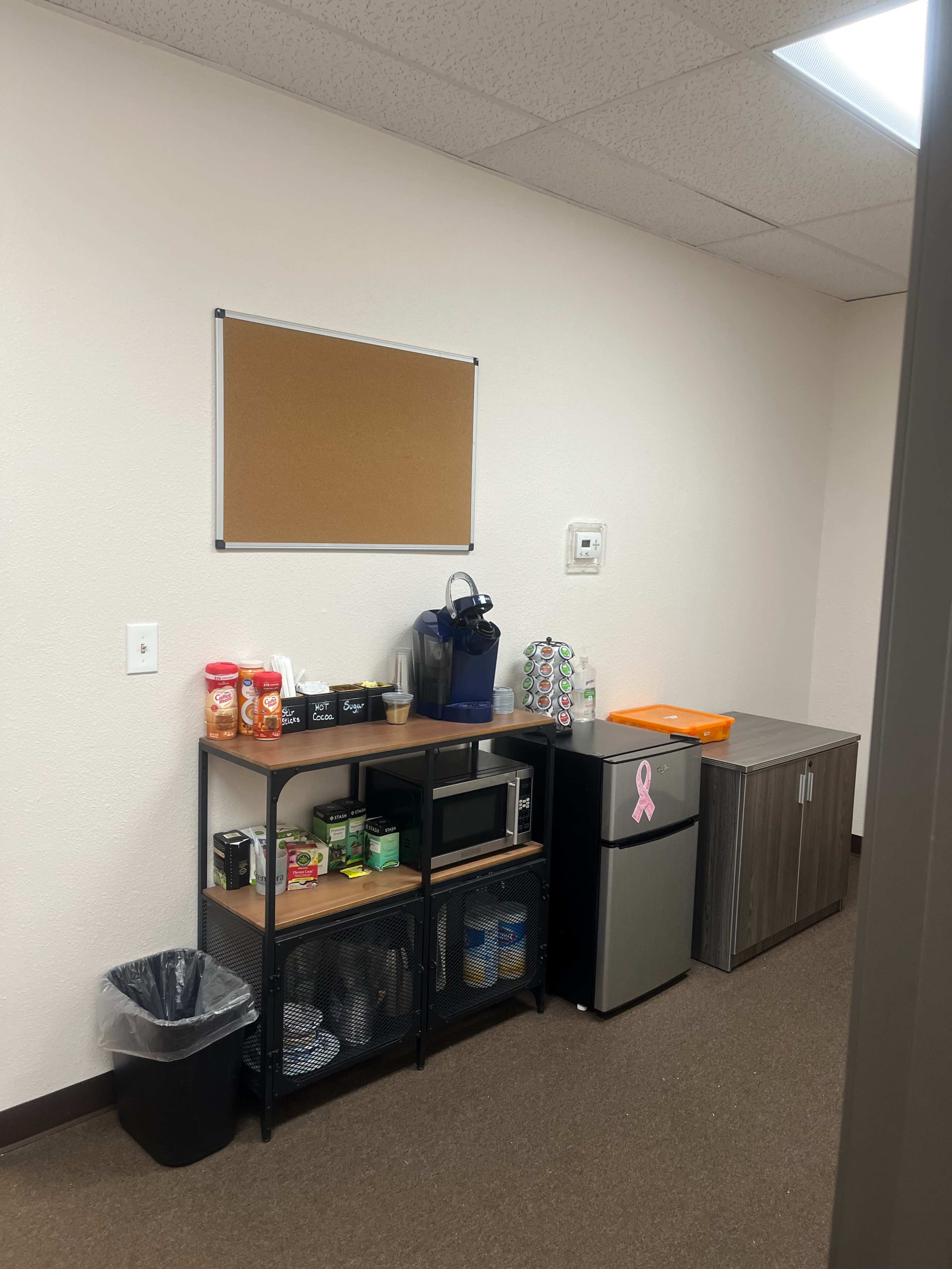 A small break room features a black shelving unit with snacks, a coffee maker, a microwave, and a mini fridge, along with a bulletin board on the wall.