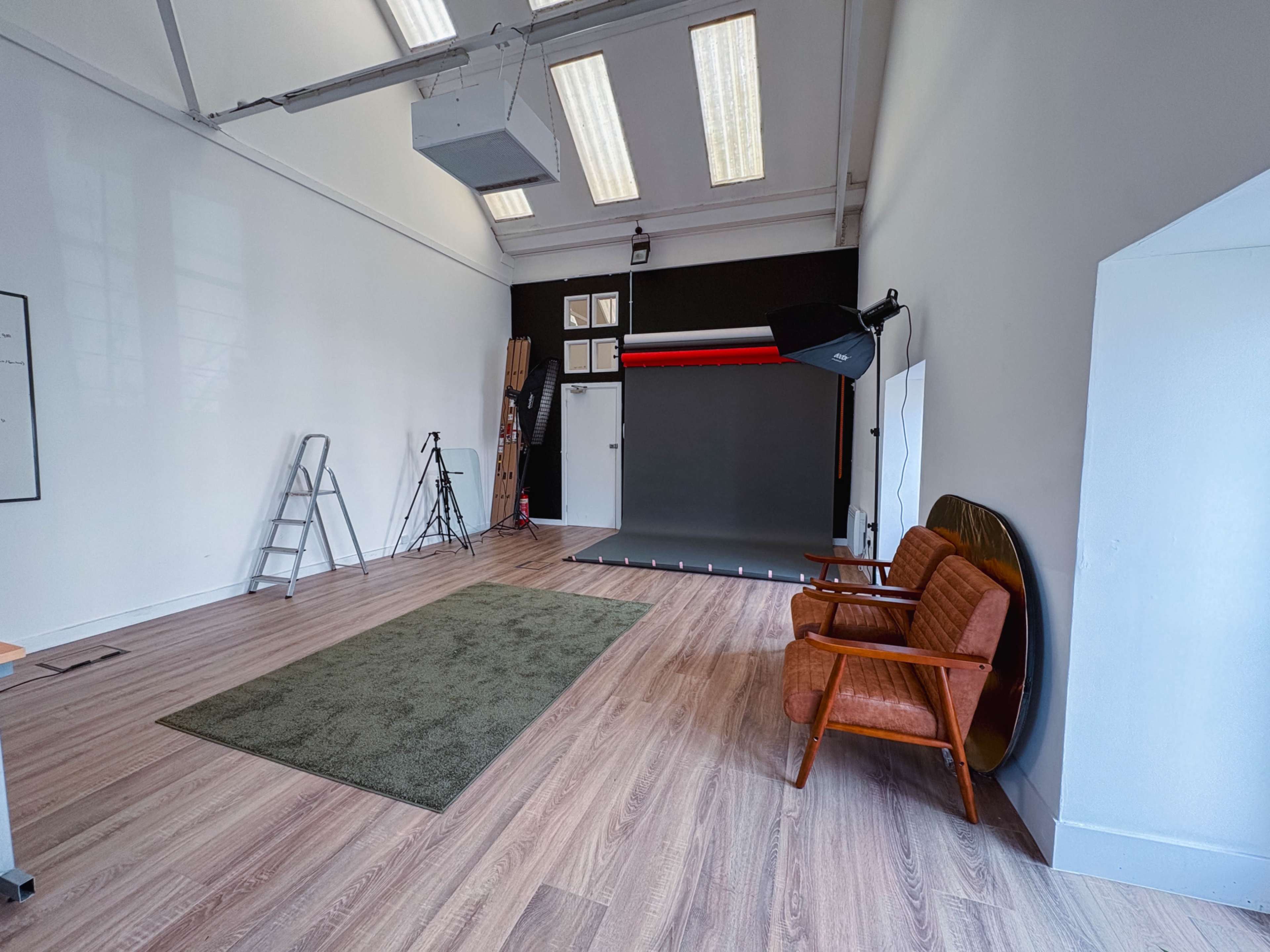 The image shows a spacious studio with wooden flooring, a green rug, a brown mid-century modern armchair, a folding ladder, and photography equipment in a bright, minimalist environment.