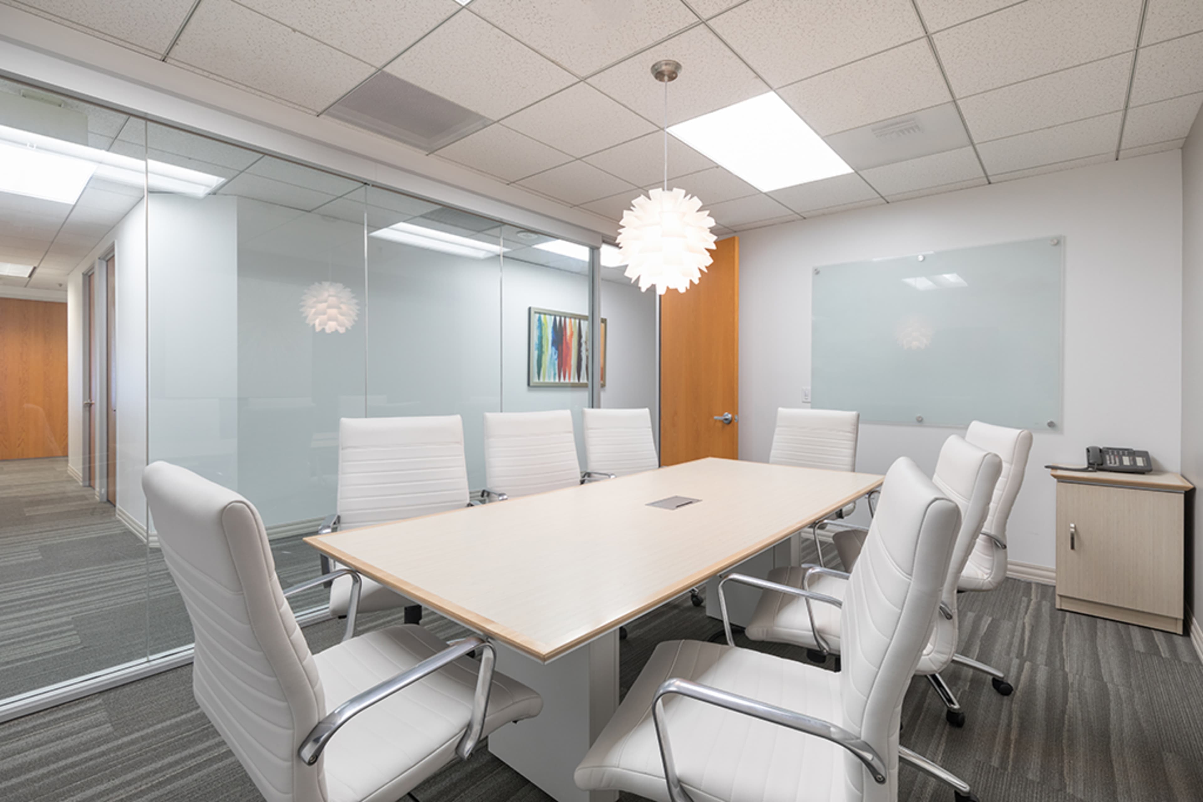 A modern conference room features a large rectangular table surrounded by white leather chairs, with glass walls and a pendant light overhead.
