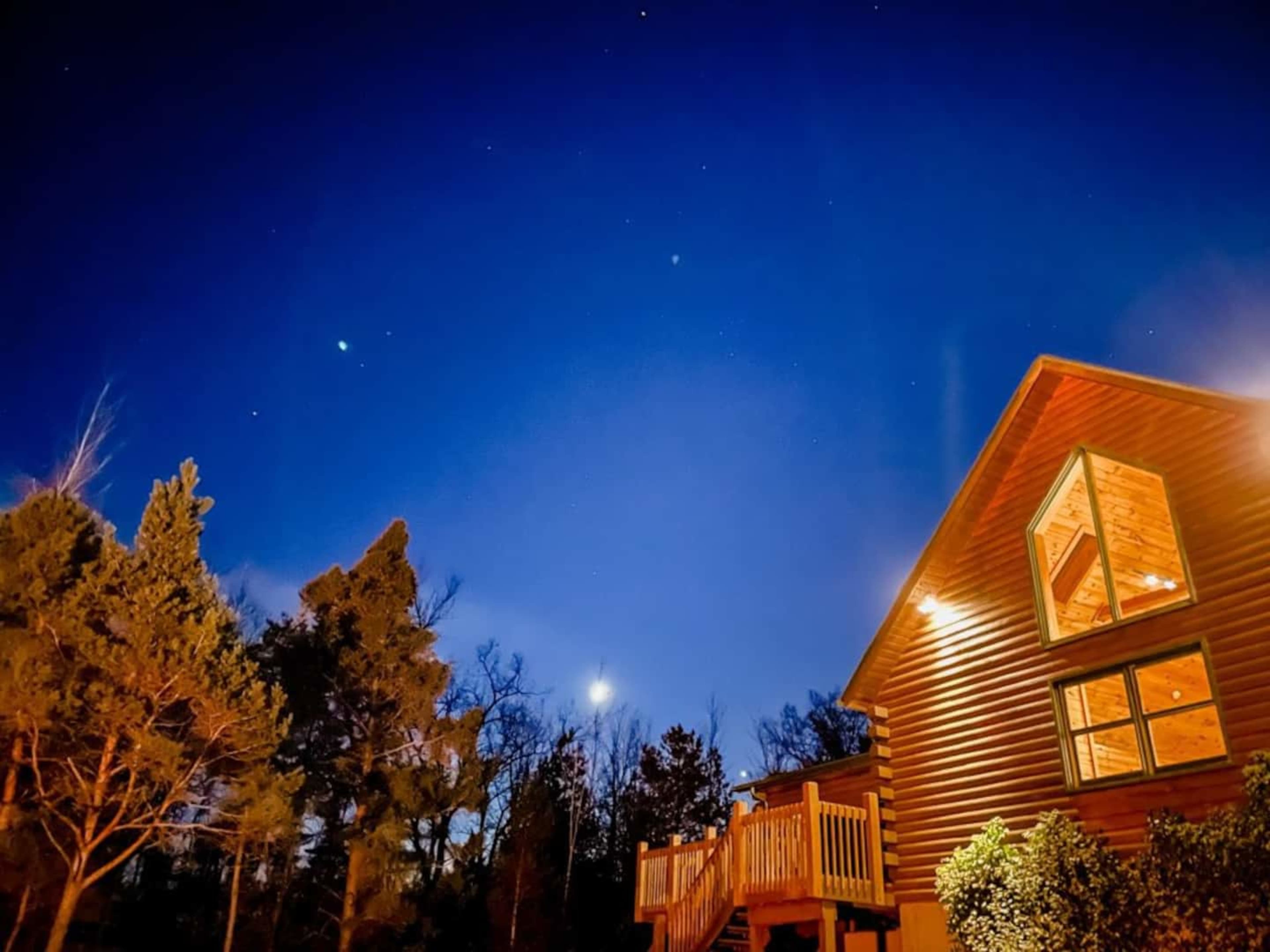 A wooden cabin is illuminated under a night sky filled with stars and a visible moon.