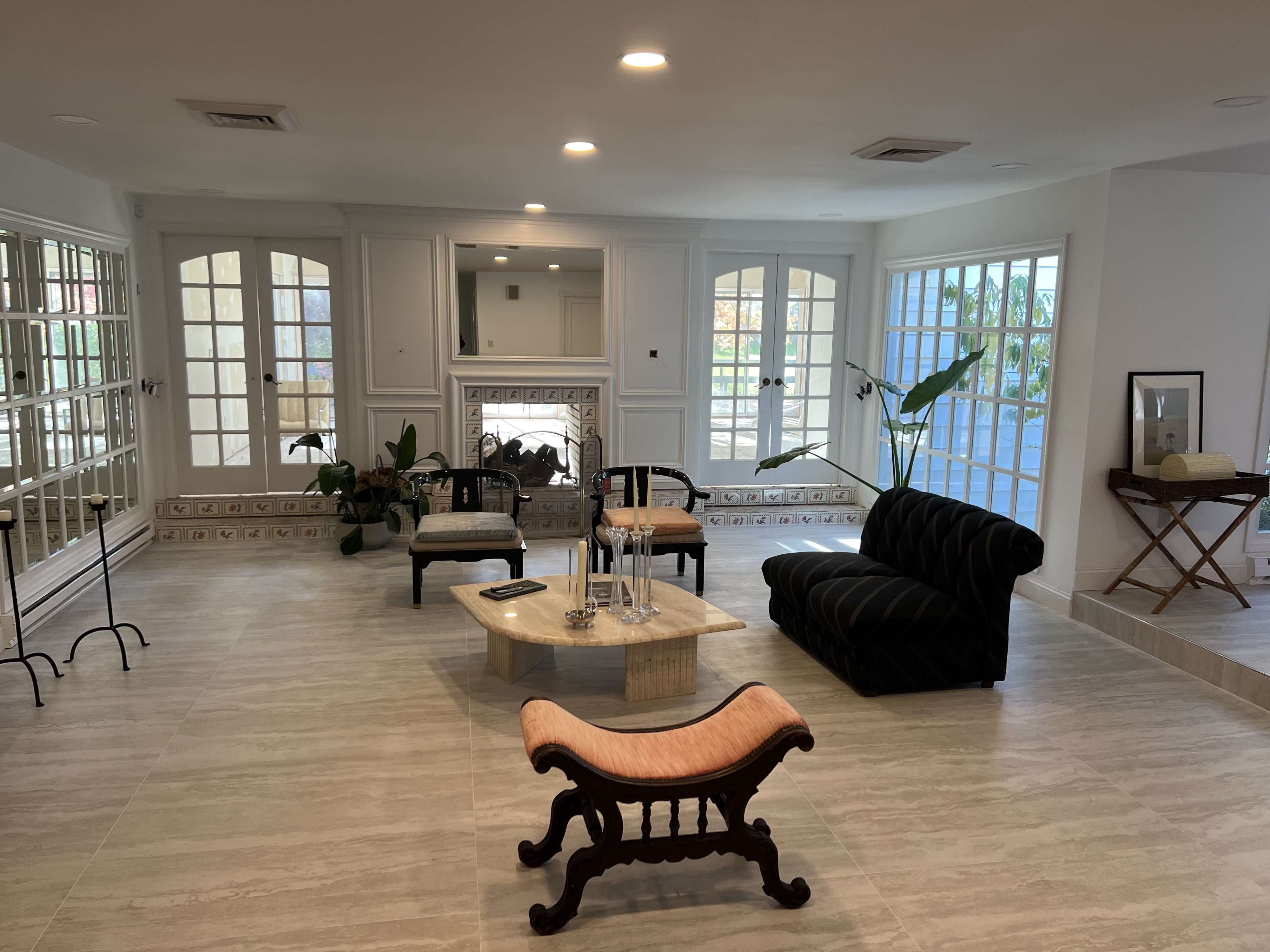 A spacious living room with large windows, a marble coffee table, a black couch, and a wooden bench, surrounded by light-colored tile flooring.