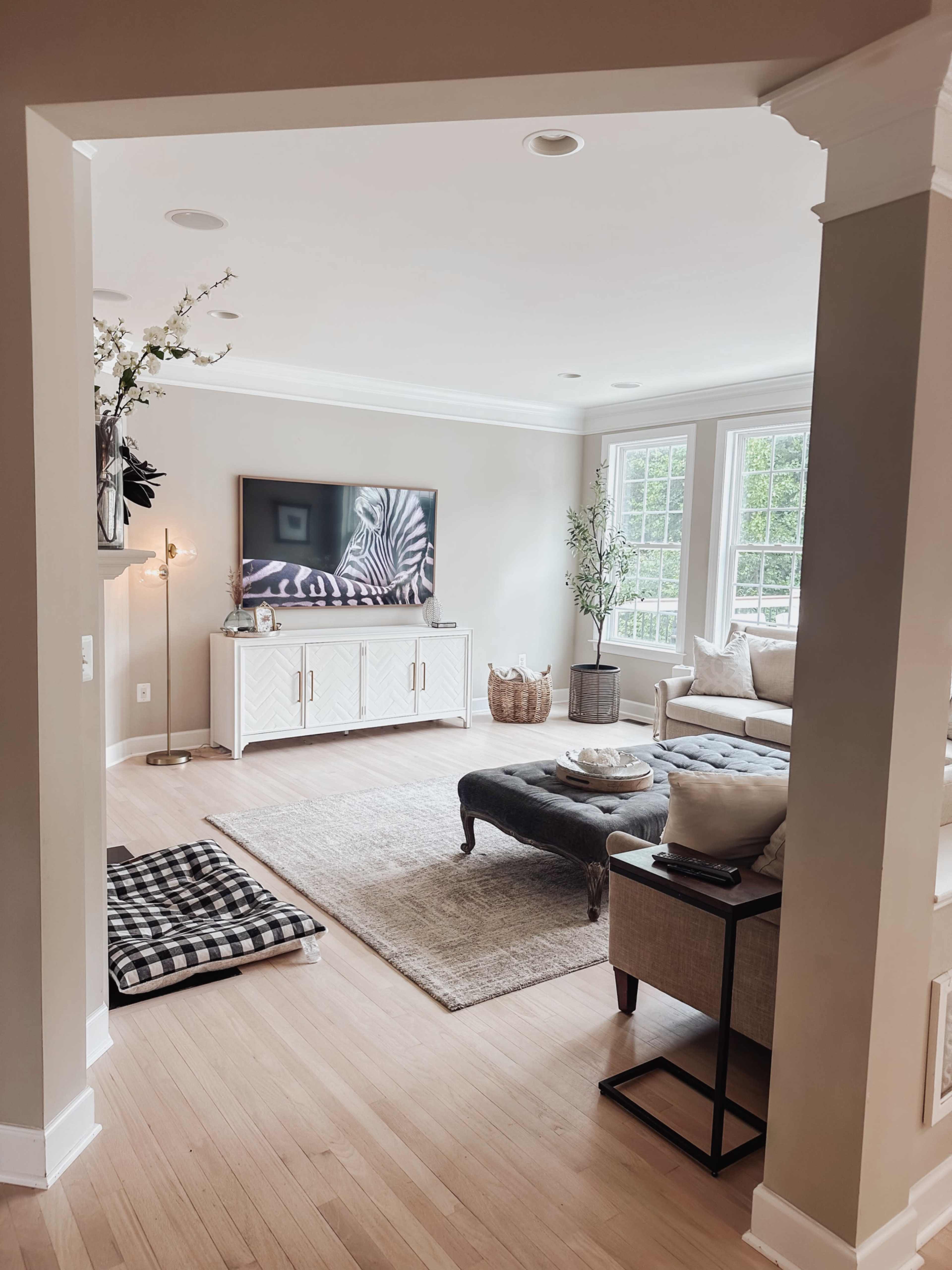 A spacious living room with a television mounted on the wall, a light-colored sofa, a coffee table, and large windows allowing natural light.