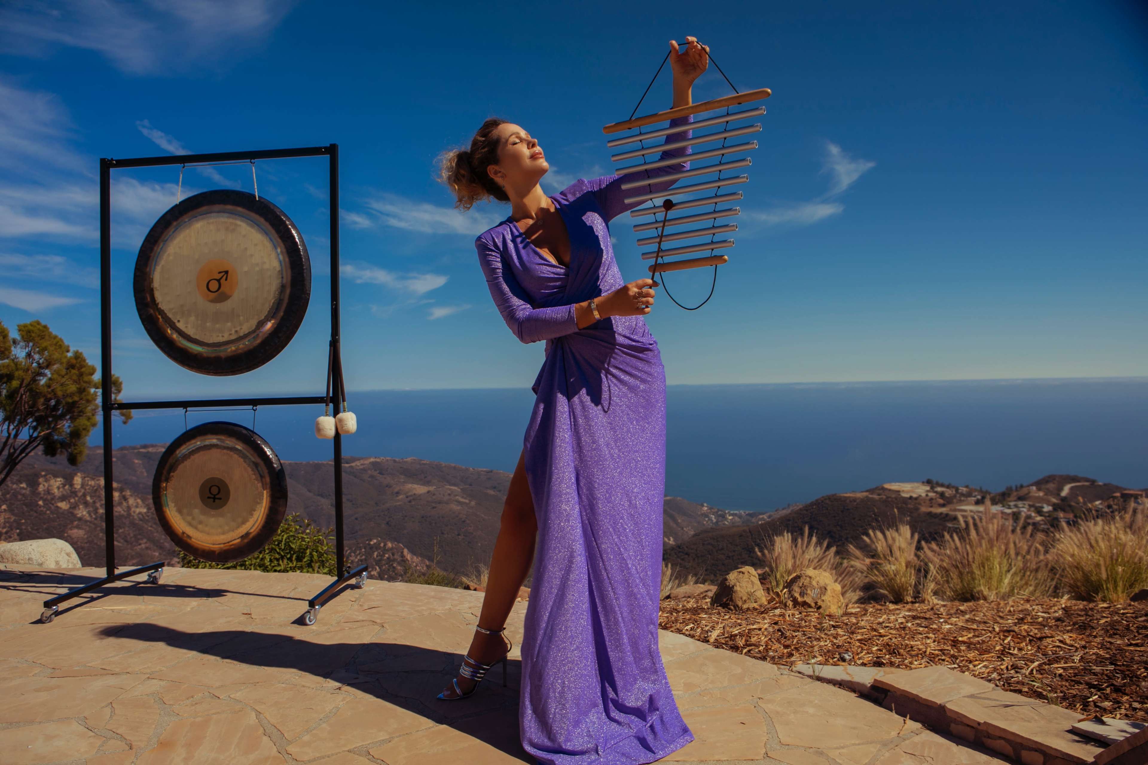 A woman in a purple dress plays a wooden instrument near two gongs with a coastal view in the background.