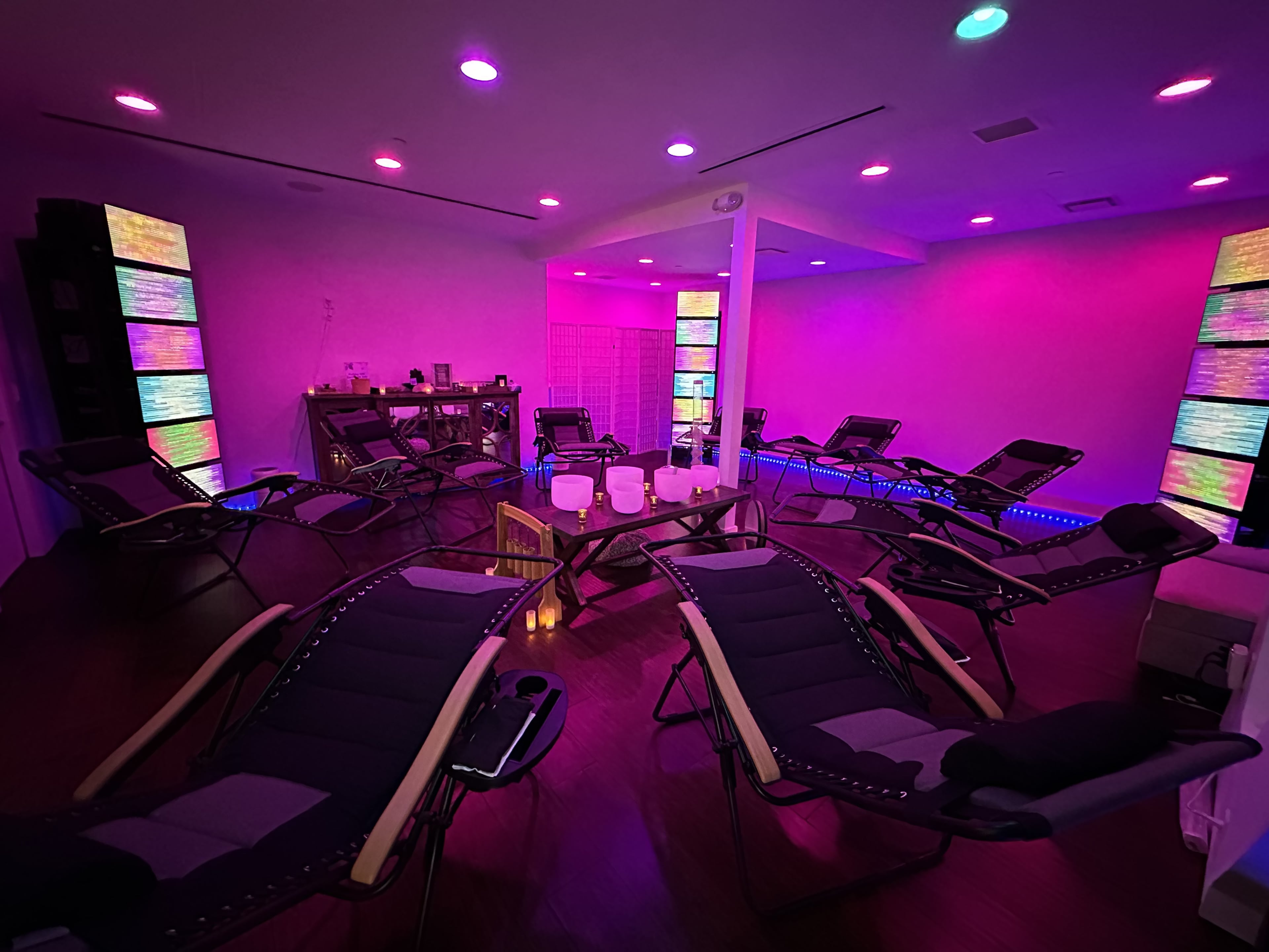 A well-lit room contains several reclining chairs arranged in a circle around a central table, with colorful light panels illuminating the walls.