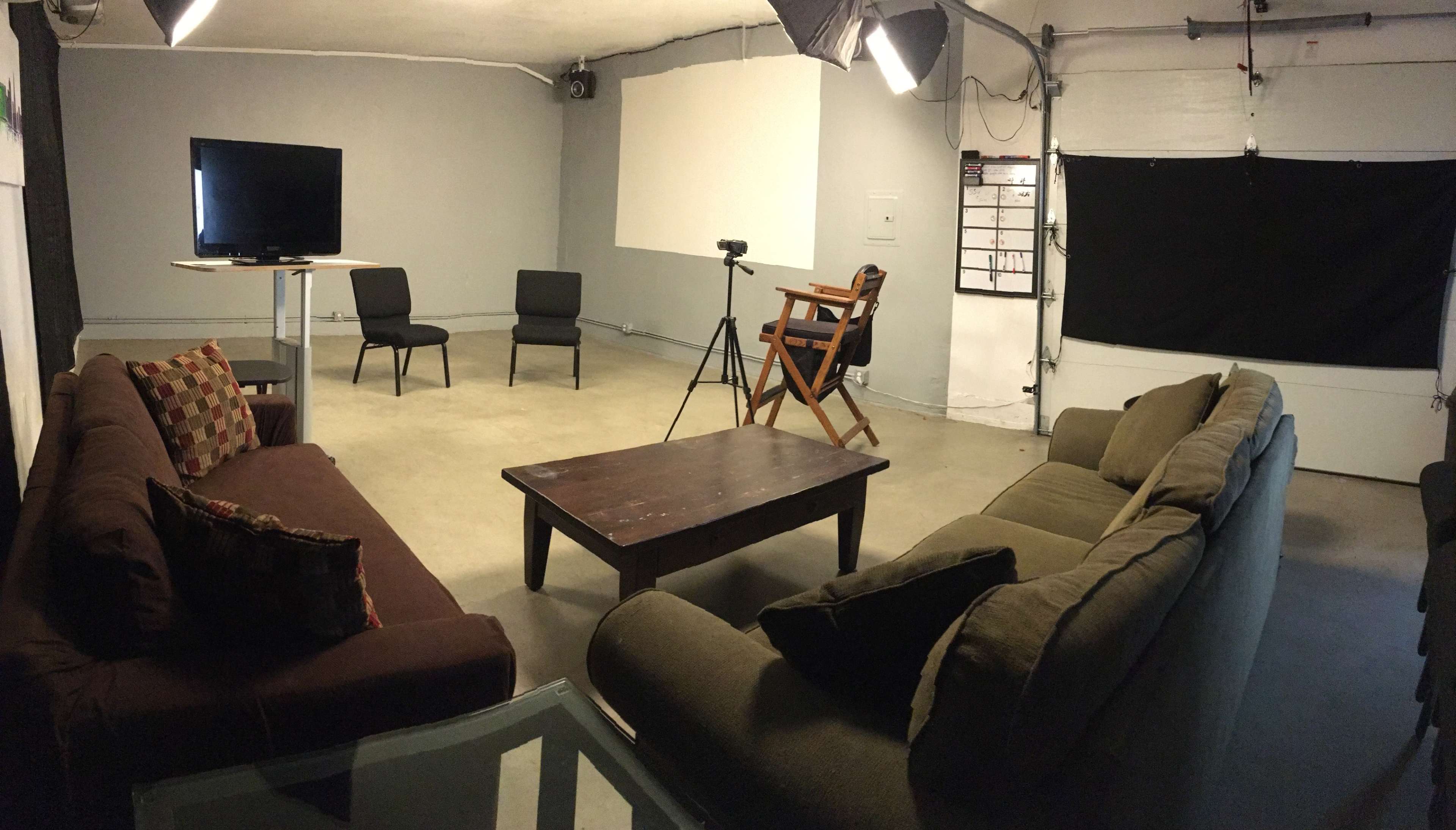 A sparse studio setup with two chairs, a table, a television, and professional lighting equipment in a neutral-colored room.