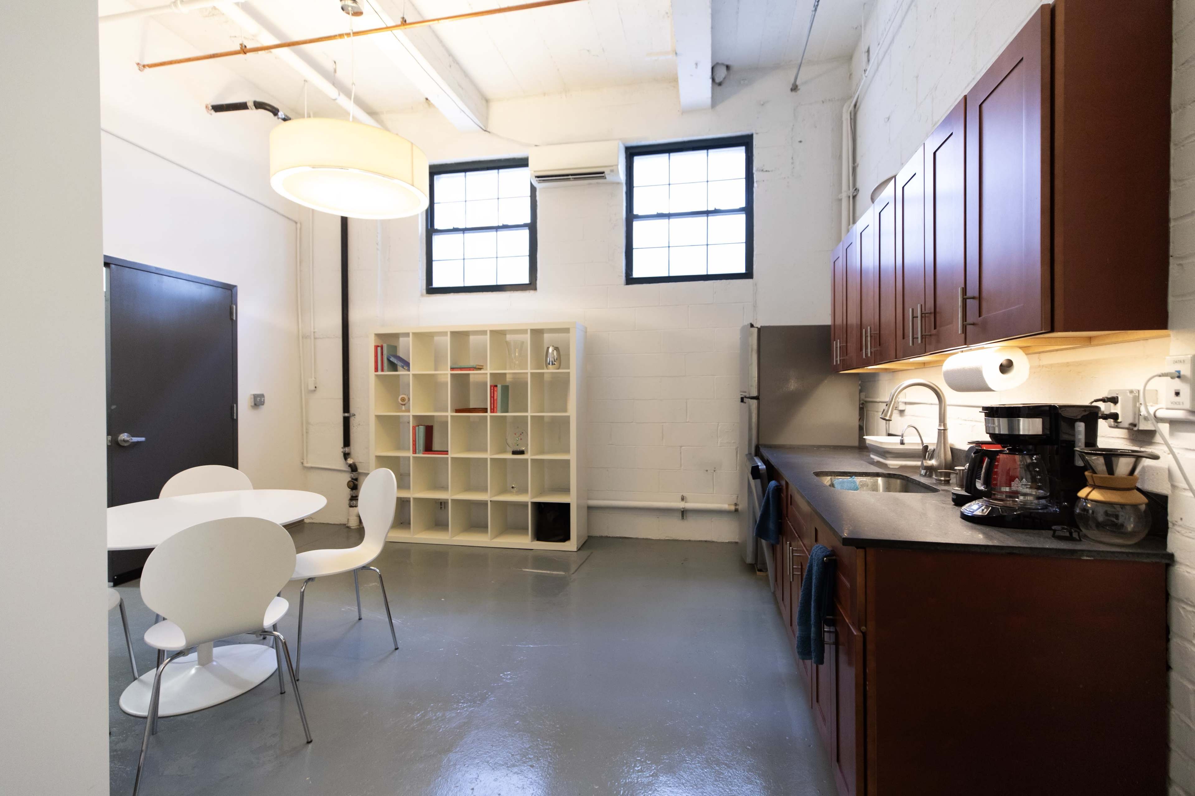 The image shows a small kitchen area featuring wooden cabinetry, a stainless steel sink, and a dining table with white chairs alongside a bookshelf and large windows.