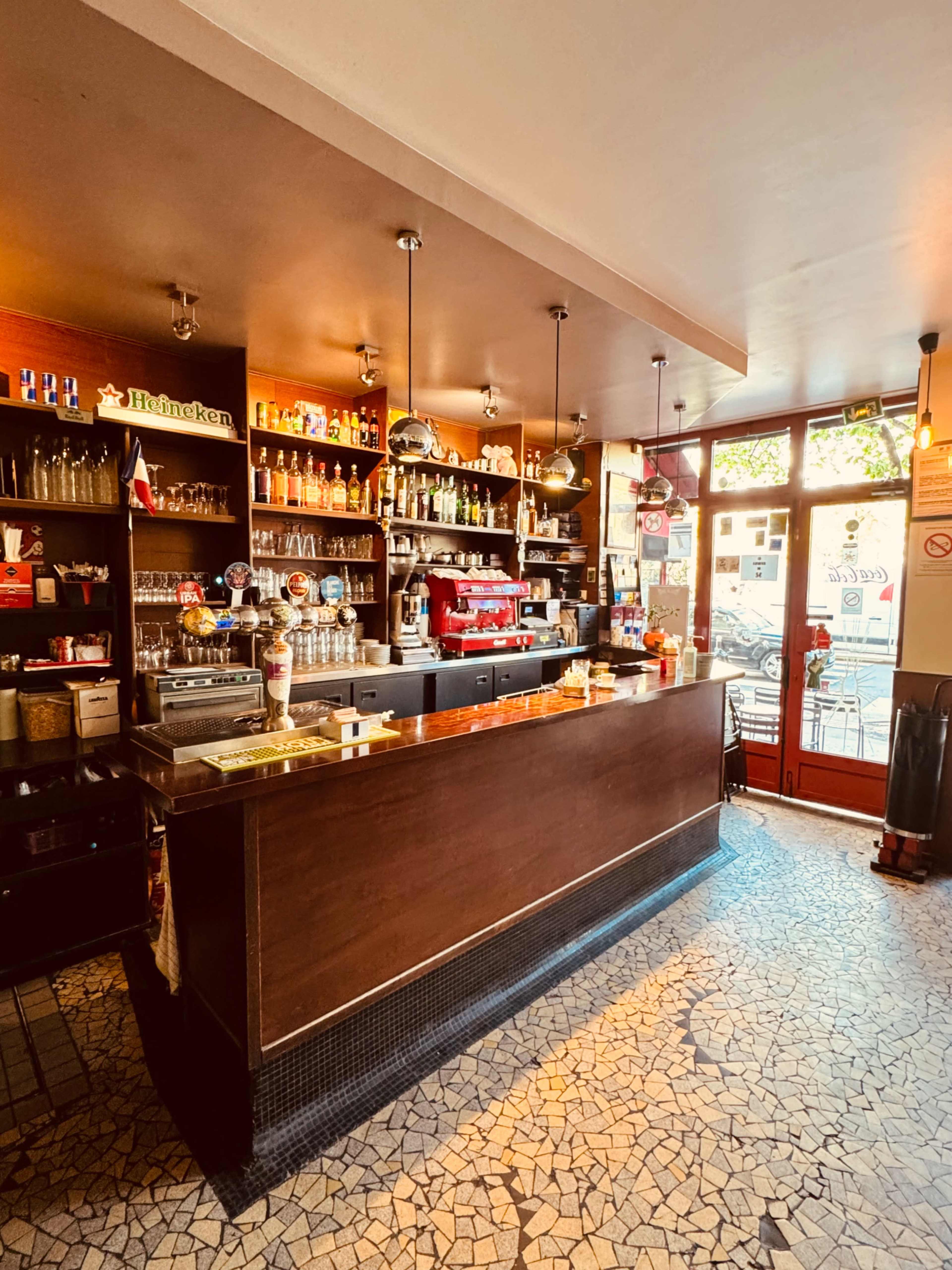 The image shows a well-stocked bar with an espresso machine, shelves of various liquor bottles, and a mosaic tile floor in a brightly lit café.