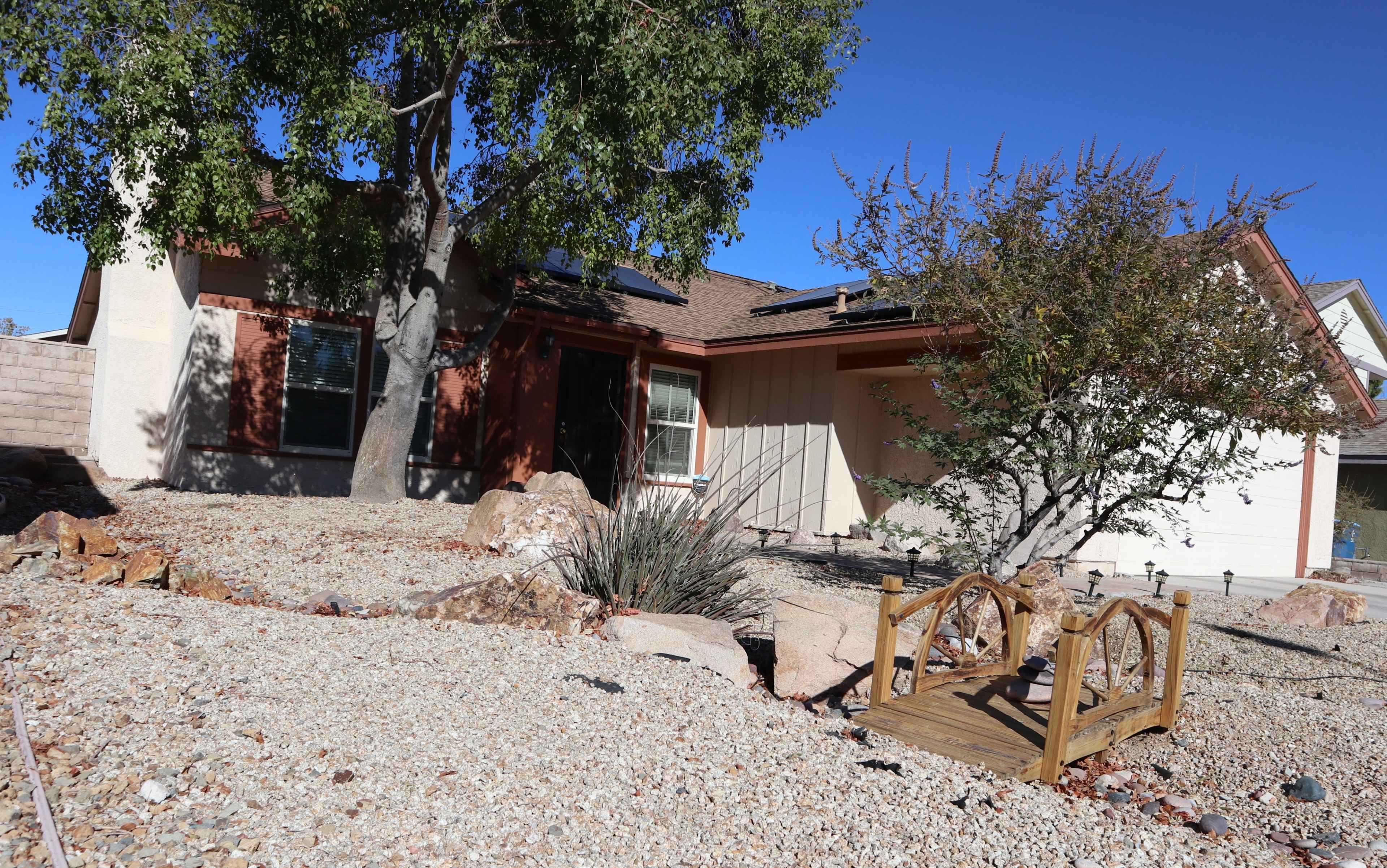A single-story house with a gravel yard, surrounded by rocks, shrubs, and solar panels on the roof.