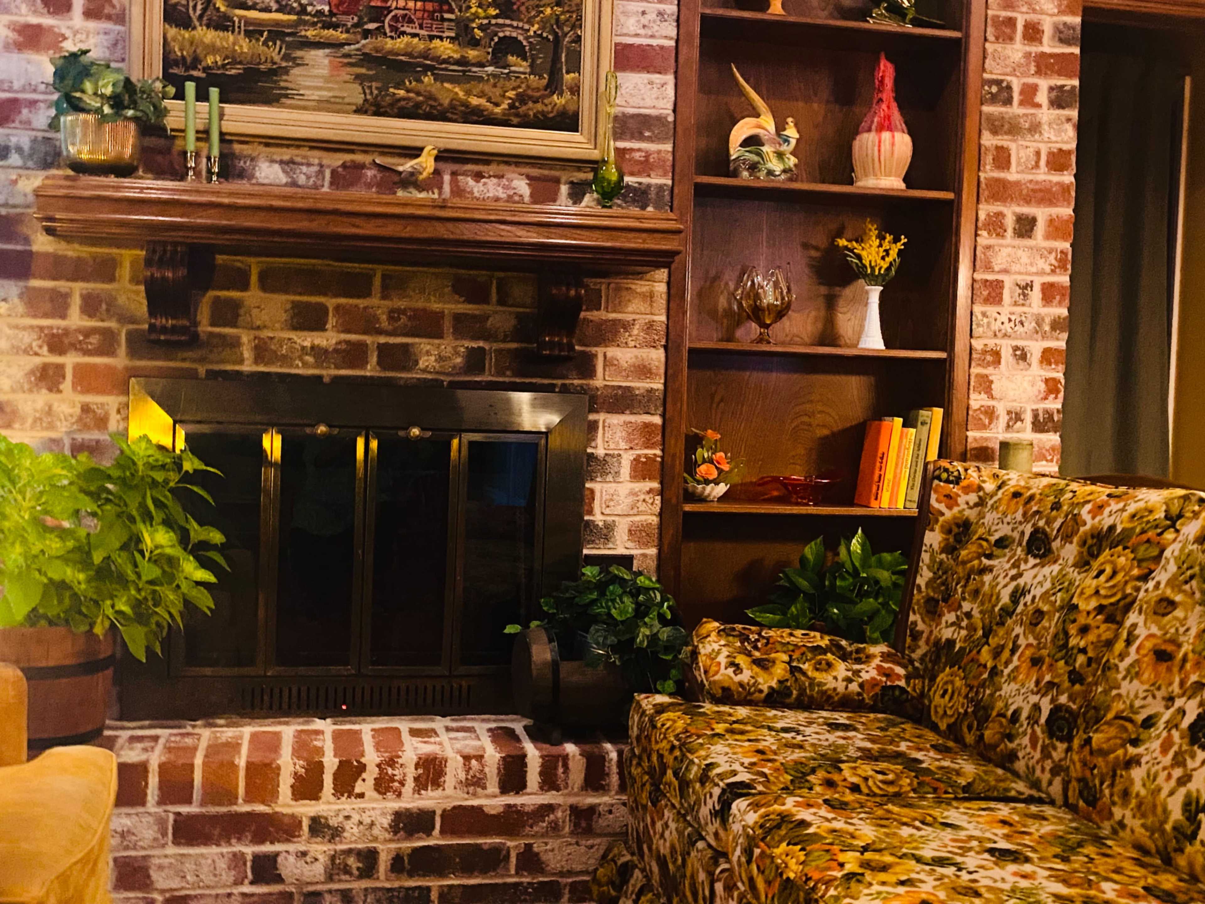 The image features a rustic living room with a brick fireplace, a floral sofa, and shelves displaying decorative items and plants.