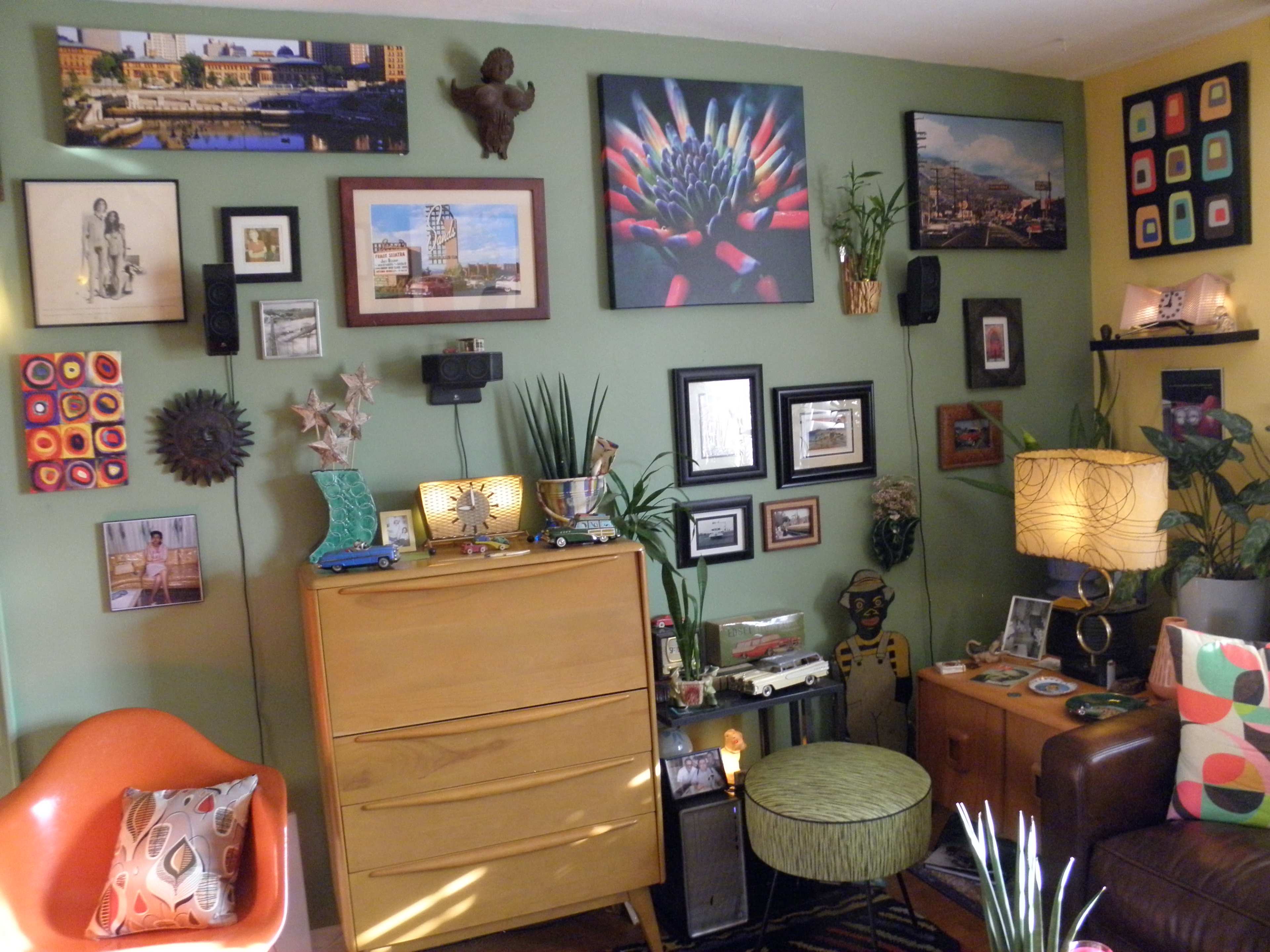 A cozy living room corner with a green wall adorned with various framed artworks, a wooden dresser, and an assortment of decorative items including plants and a lamp.