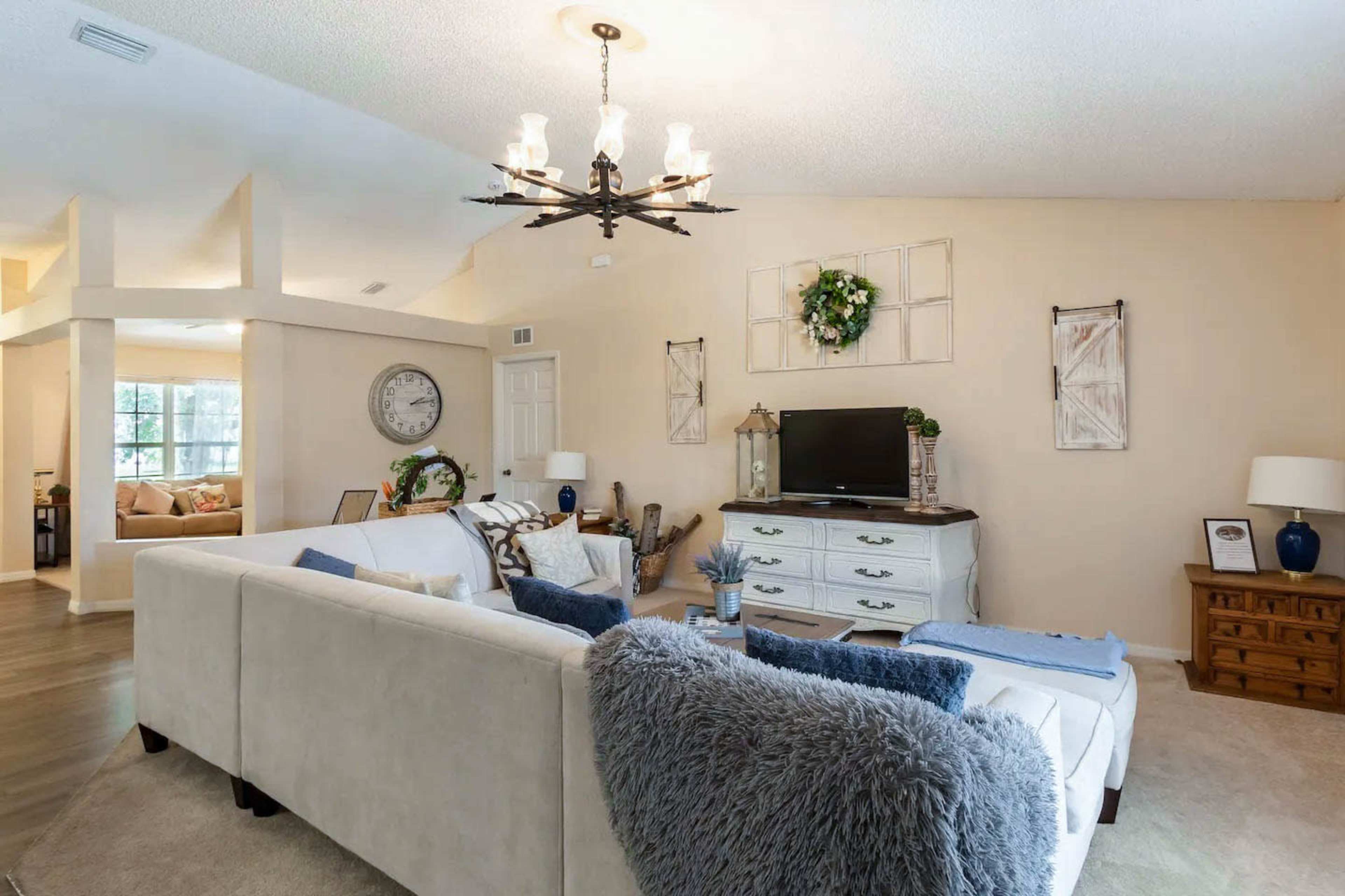 The image shows a living room featuring a white sectional sofa, a television on a light-colored cabinet, and decorative wall hangings.