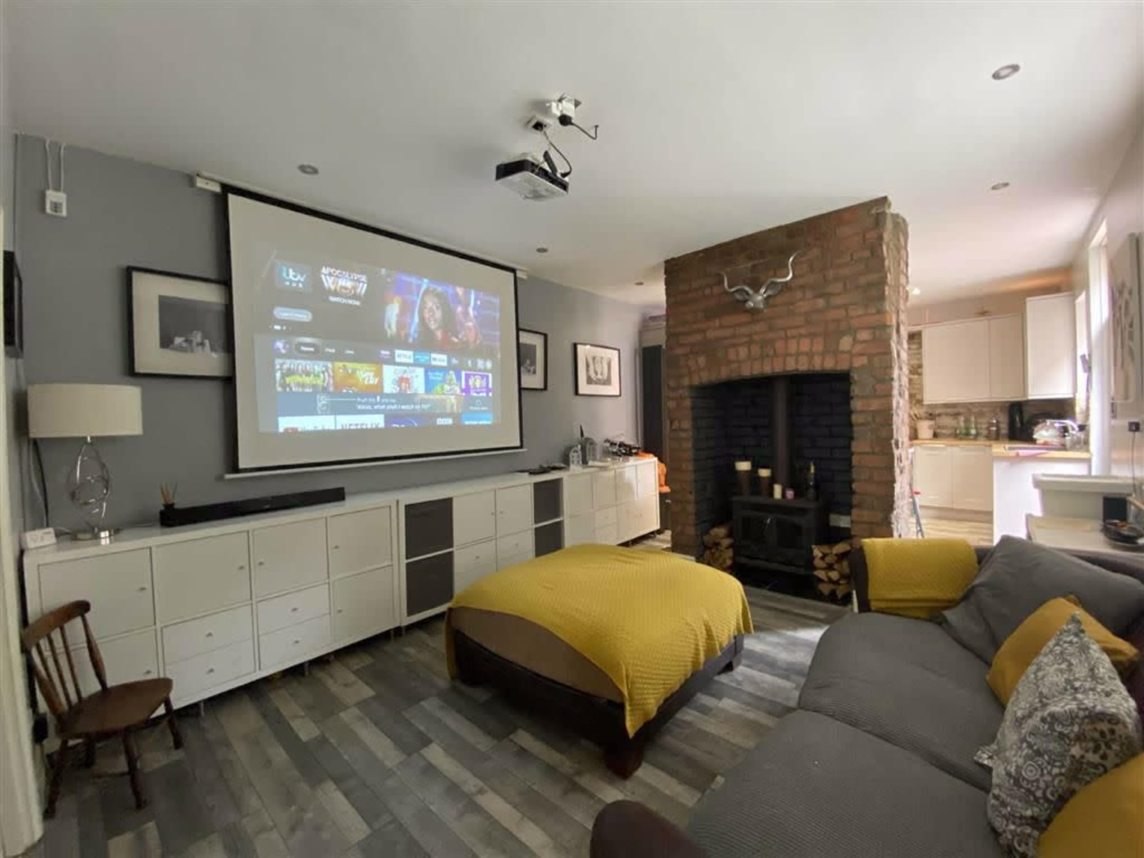 A cozy living room features a large projection screen displaying various media options, with a brick fireplace, a sofa, and a wooden chair in the space.