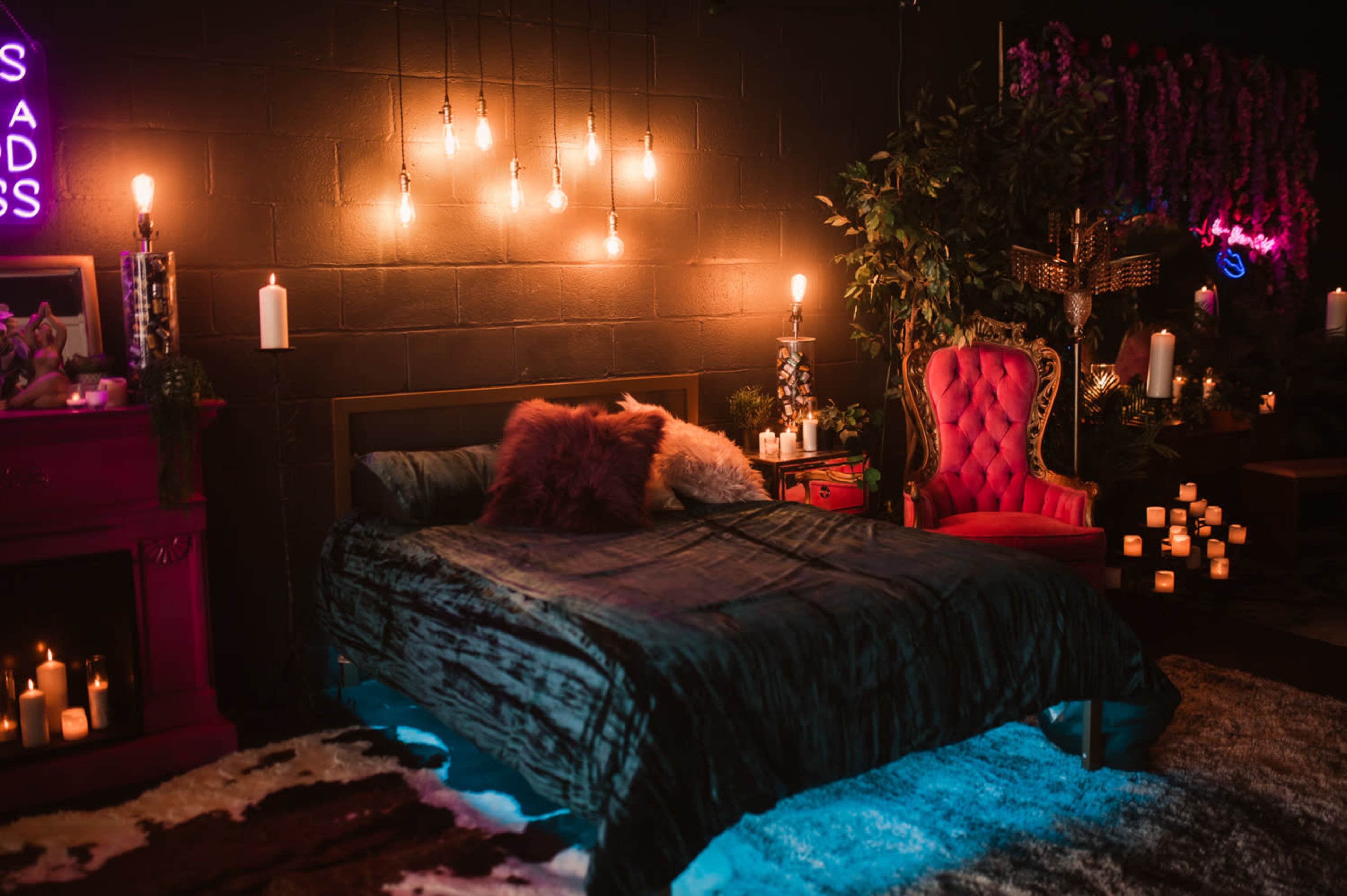 The scene features a dimly lit bedroom with a bed adorned in dark bedding, surrounded by candles and a vibrant pink armchair against a wall decorated with greenery and hanging lights.