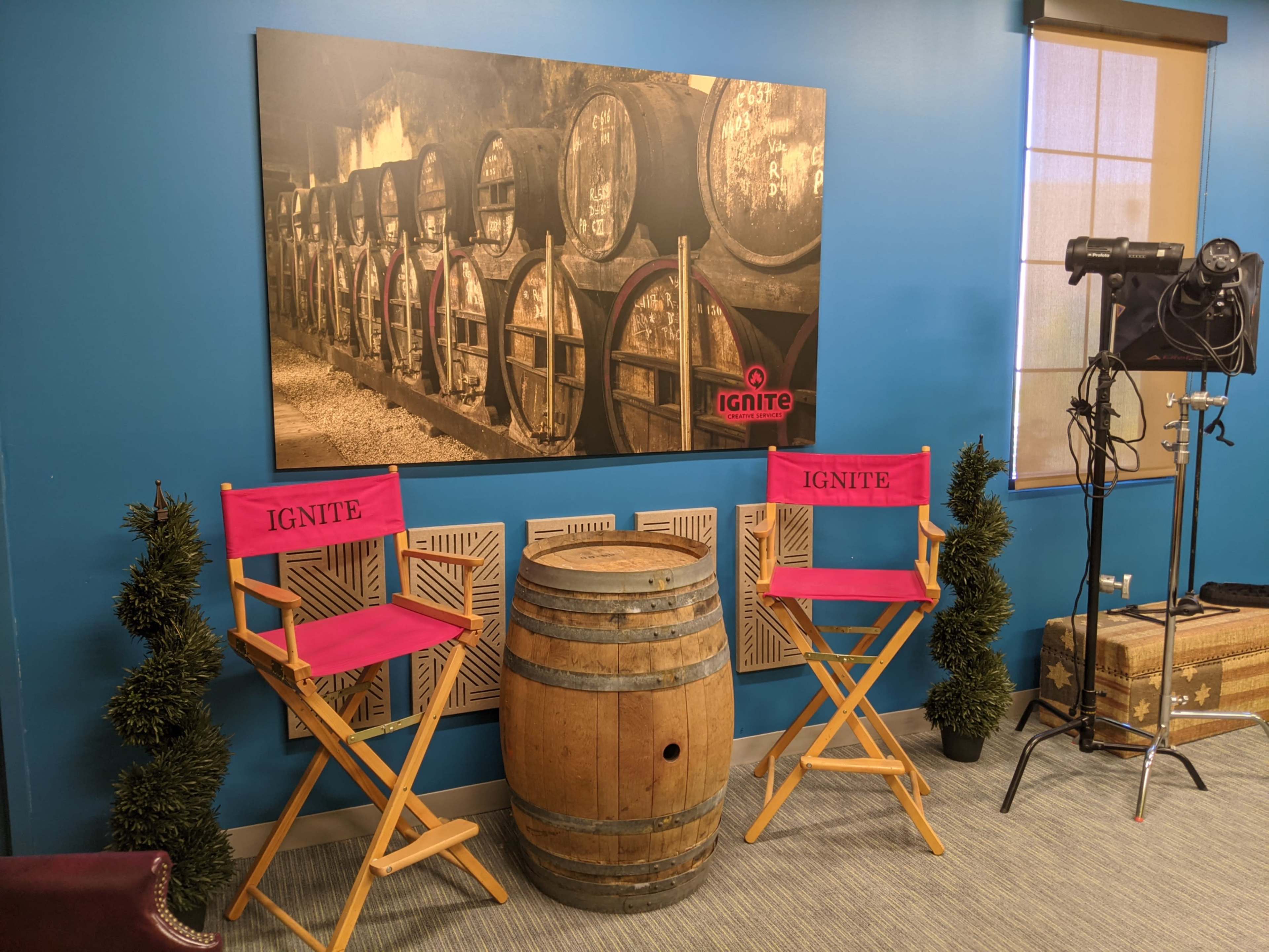 The image shows a room with two pink director's chairs labeled "IGNITE," a wooden barrel, and a large black-and-white photograph of barrels in a cellar on the wall.