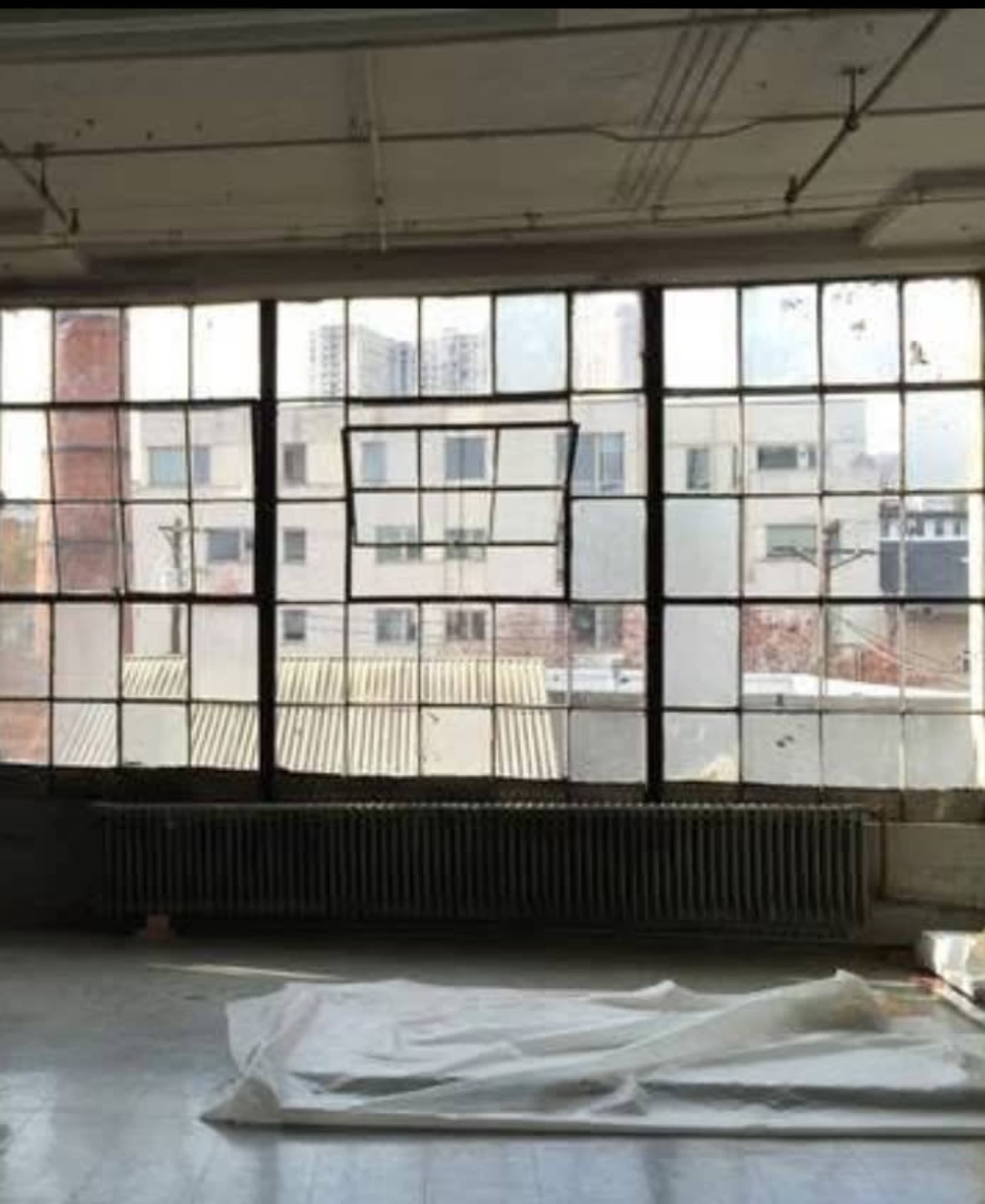 The image shows a large industrial window with multiple panes, revealing a view of neighboring buildings and a partially covered floor inside an empty room.
