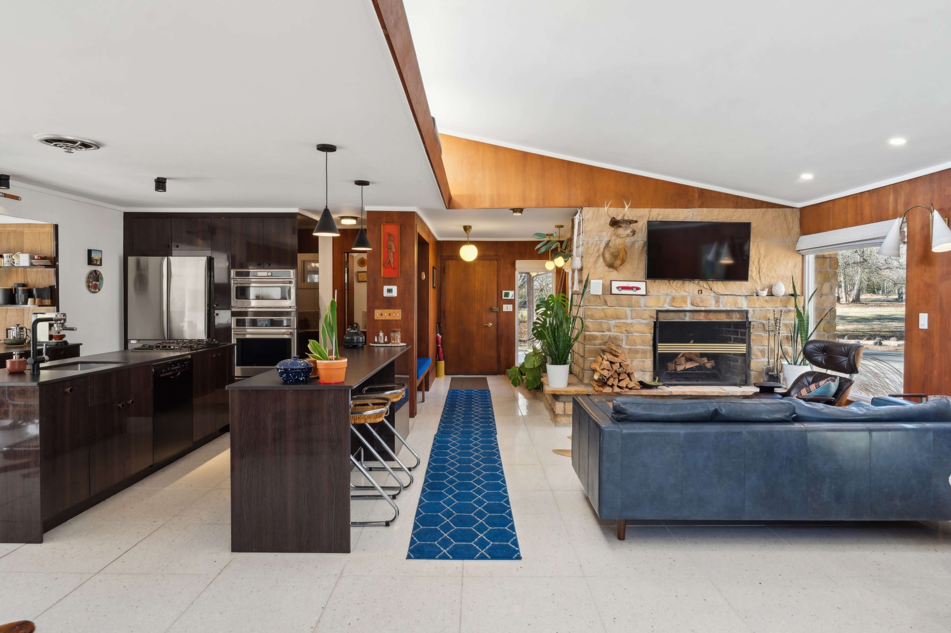 The image shows a modern open-concept living space featuring a kitchen, dining area, and lounge with a stone fireplace and large windows.