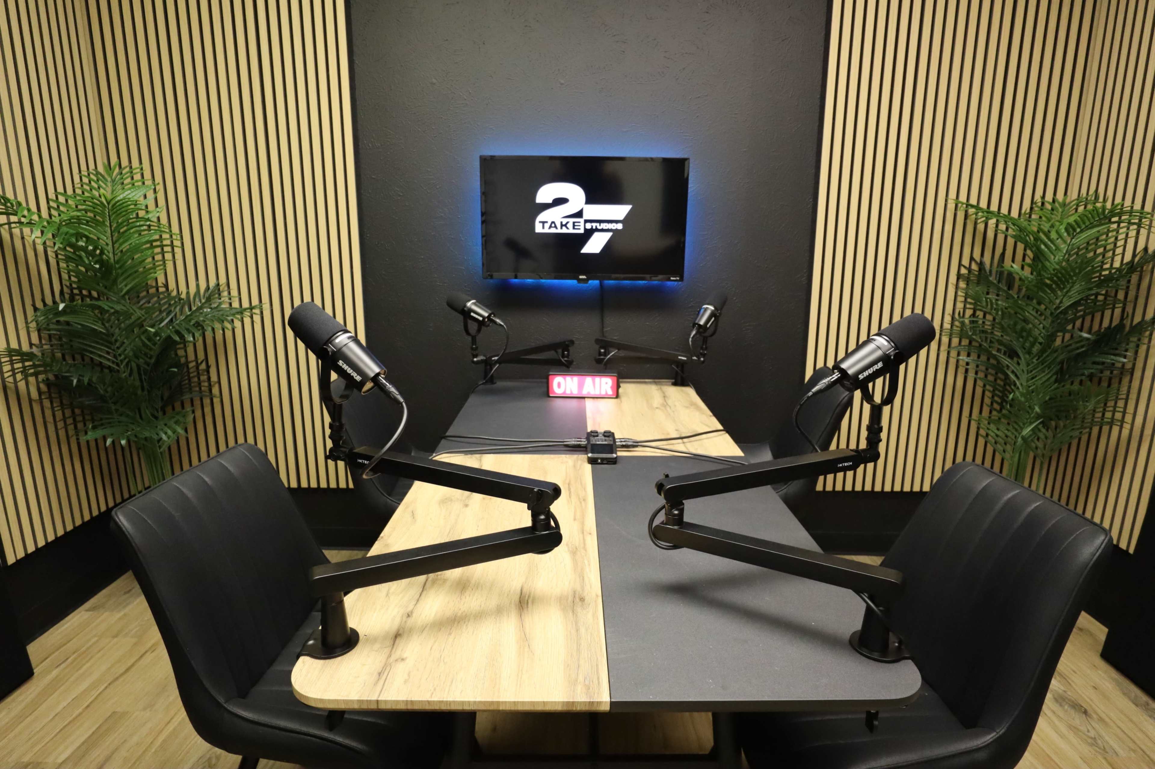 A podcast studio with two microphones positioned at an elongated wooden table, a "ON AIR" sign, and a television screen in the background.