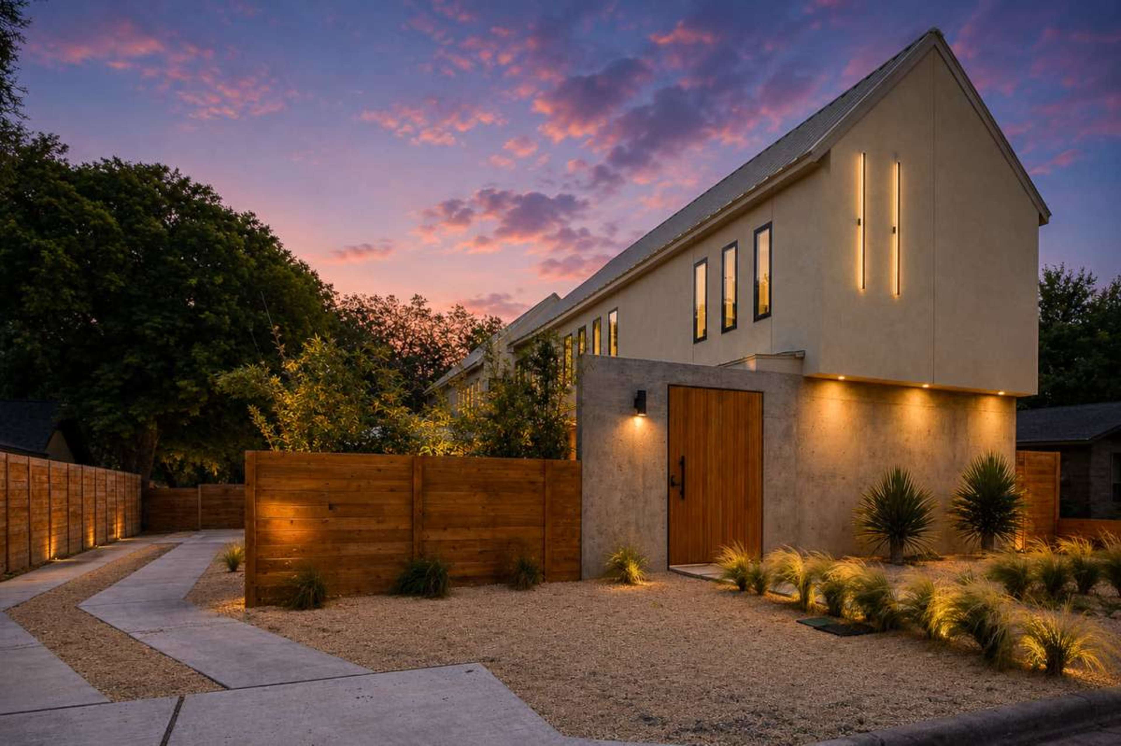 The modern house features a wooden door and sleek exterior lighting, set against a twilight sky with colorful clouds.