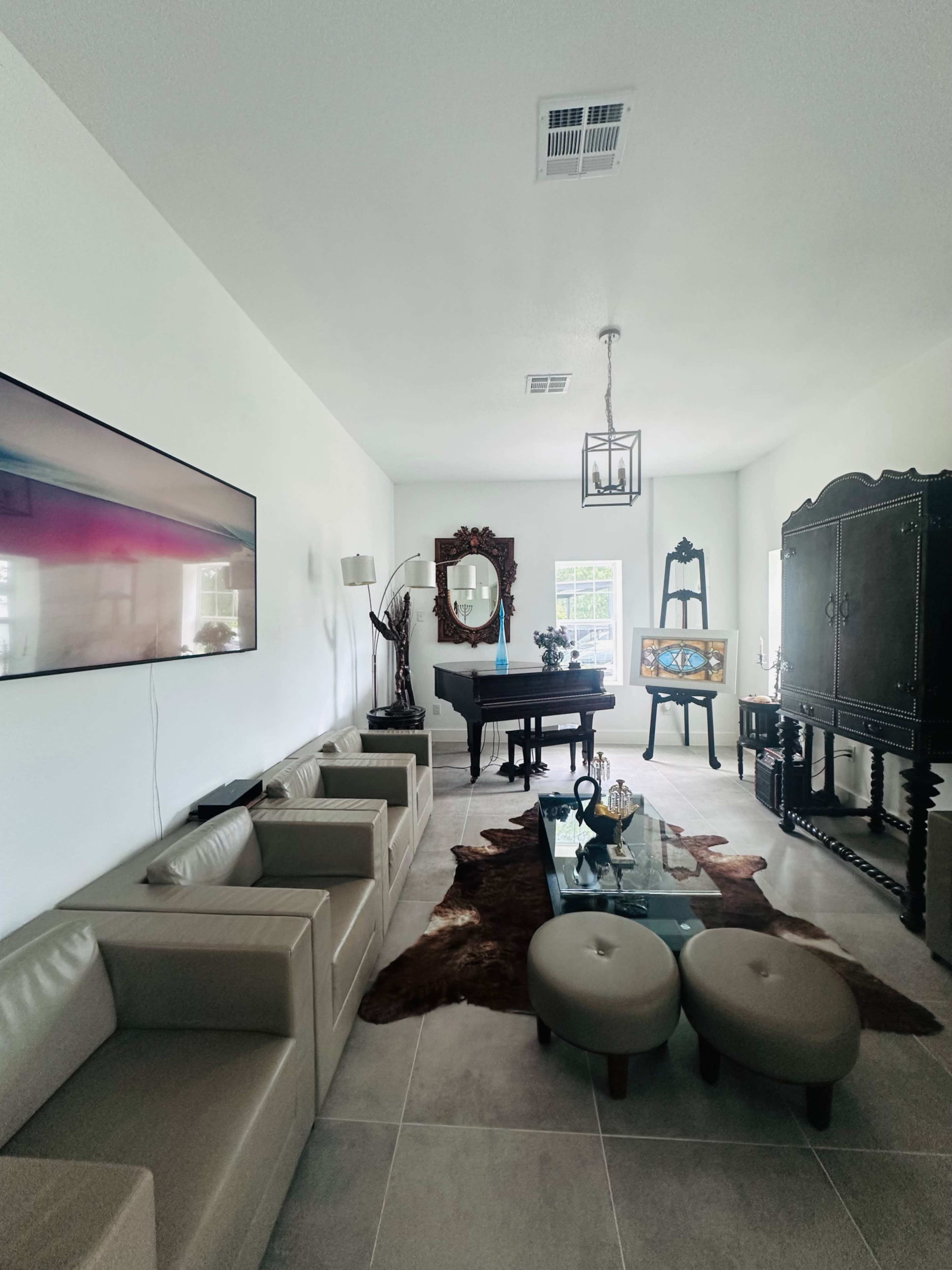 The image shows a well-lit living room featuring a modern sofa, a glass coffee table, and various decorative elements, including a piano and a large wooden cabinet.