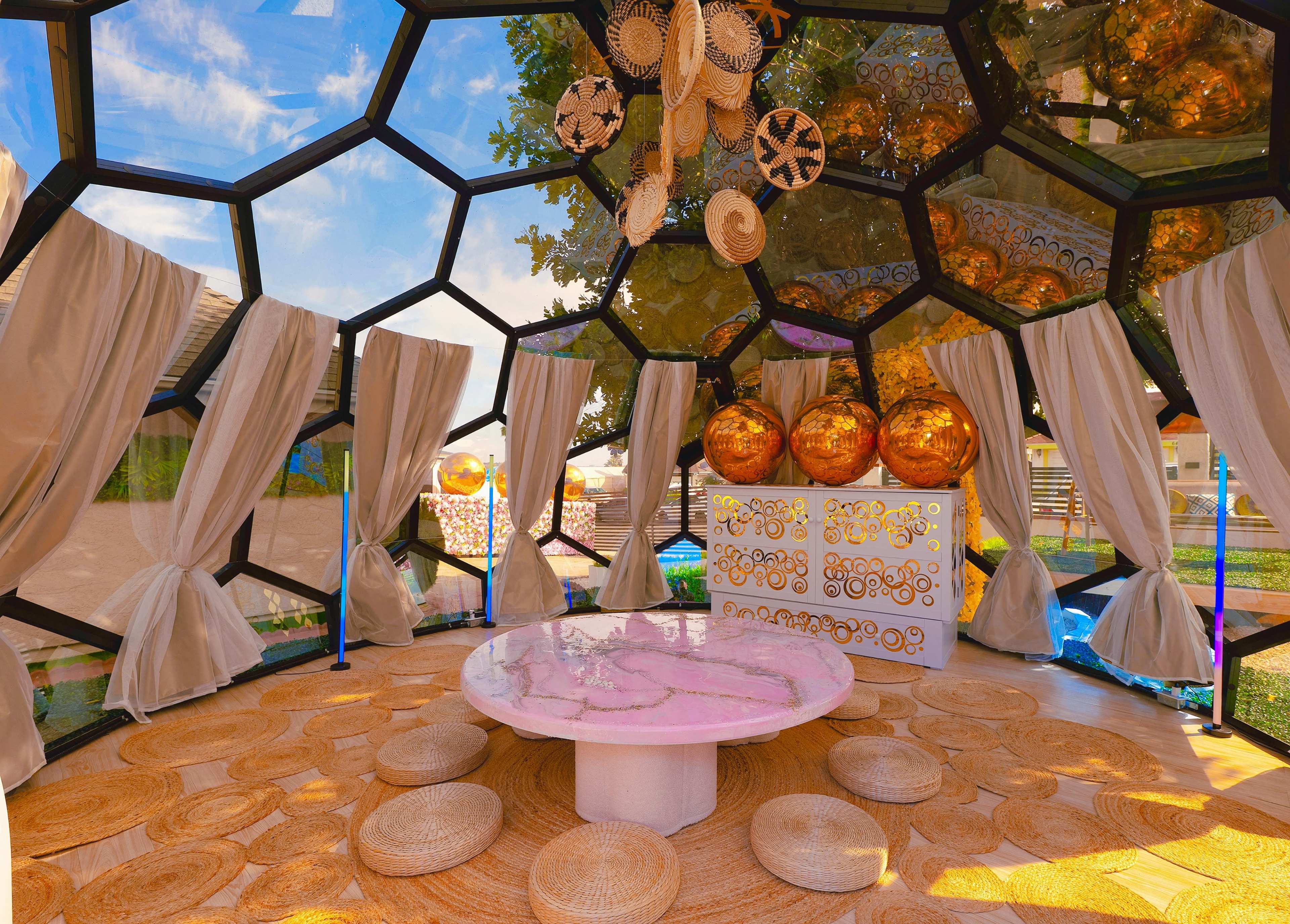 The image features a modern, geodesic dome interior with a circular table, decorative hanging items, and natural light filtering through large windows.