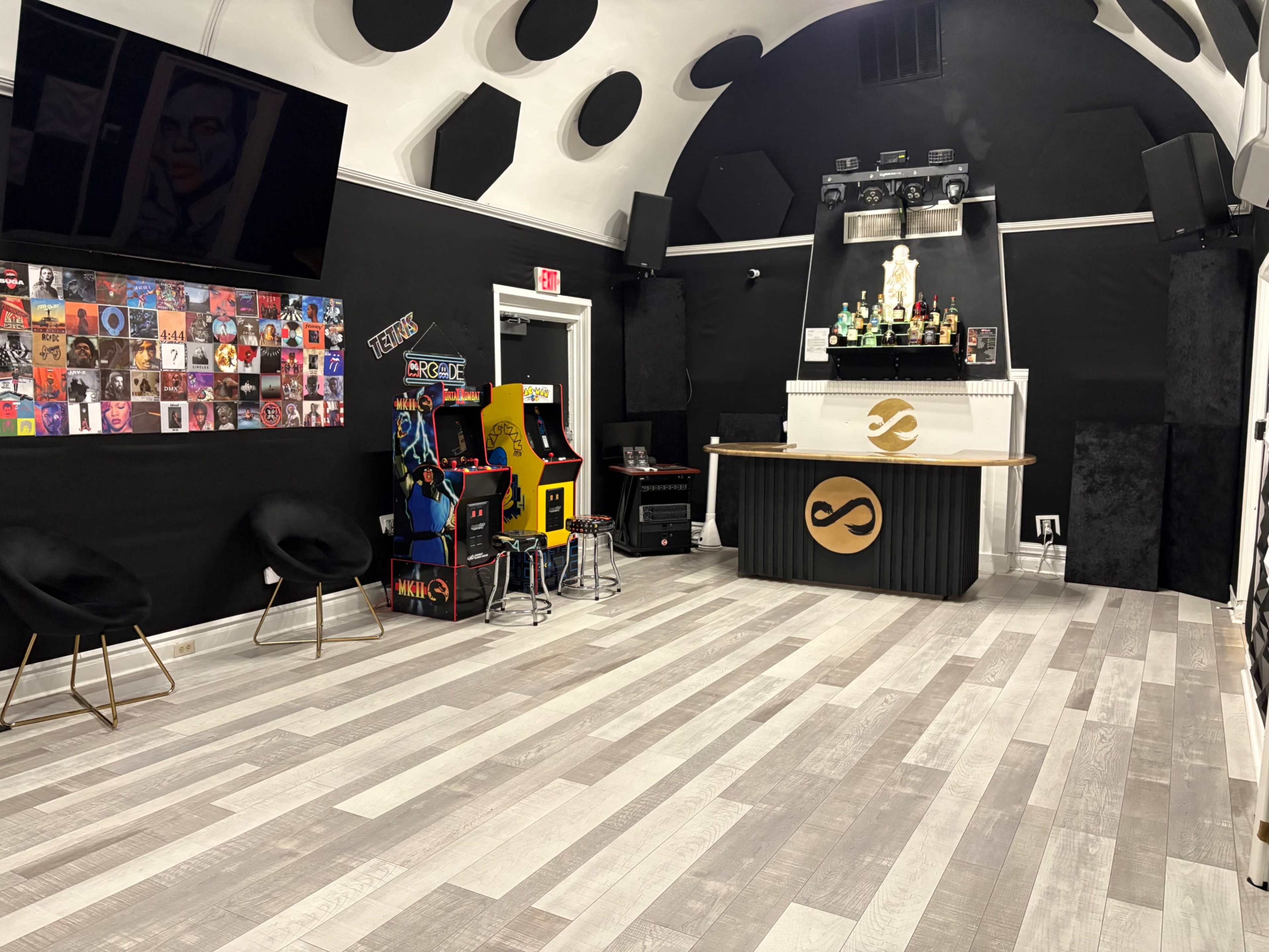 The image shows a modern lounge area with a bar, arcade games, a wall of framed artwork, and sleek black and light wood flooring.
