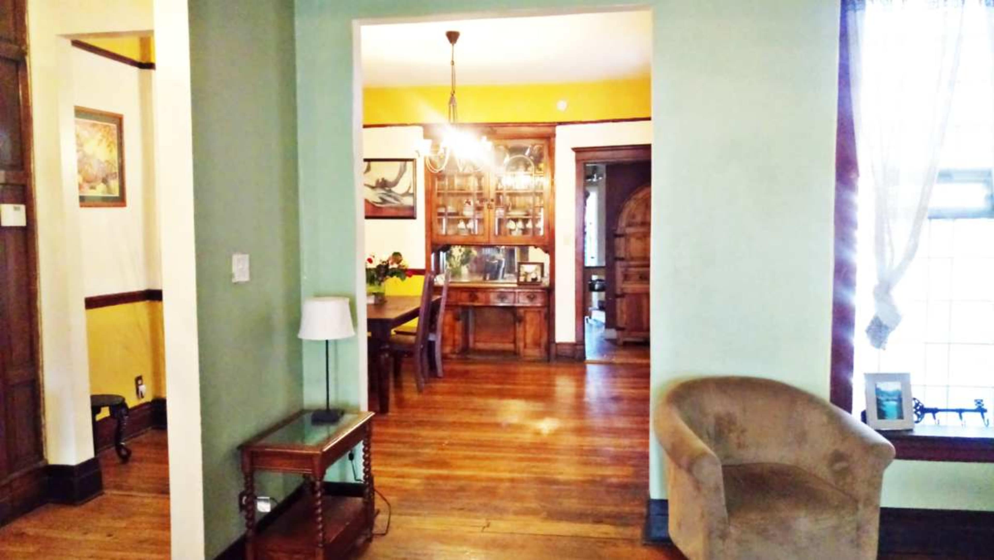 A view of a room with a light green wall leading into a dining area, featuring a wooden dining set and a chandelier.
