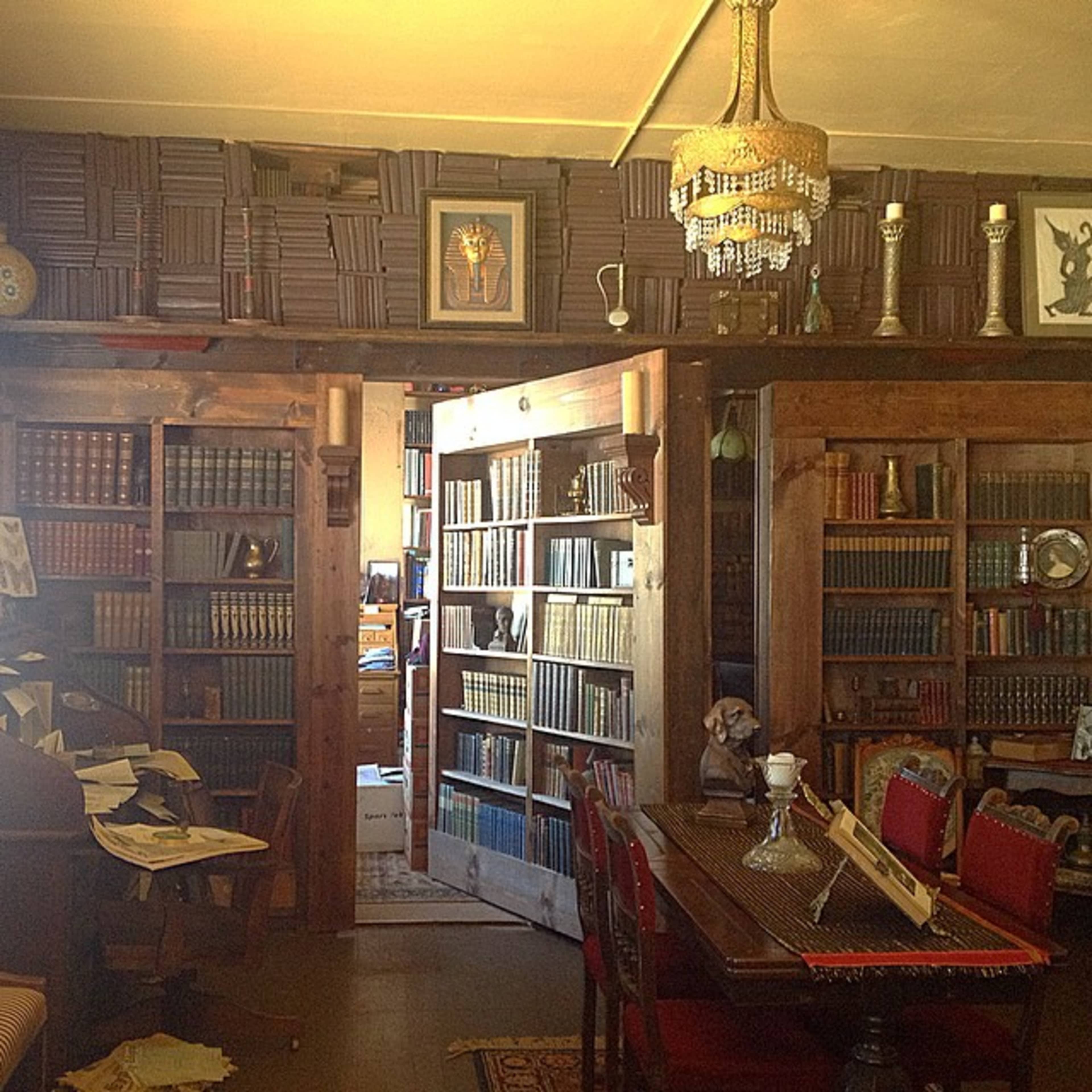 A cozy library with wooden bookshelves filled with books, a dining table, and decorative items, illuminated by a chandelier.