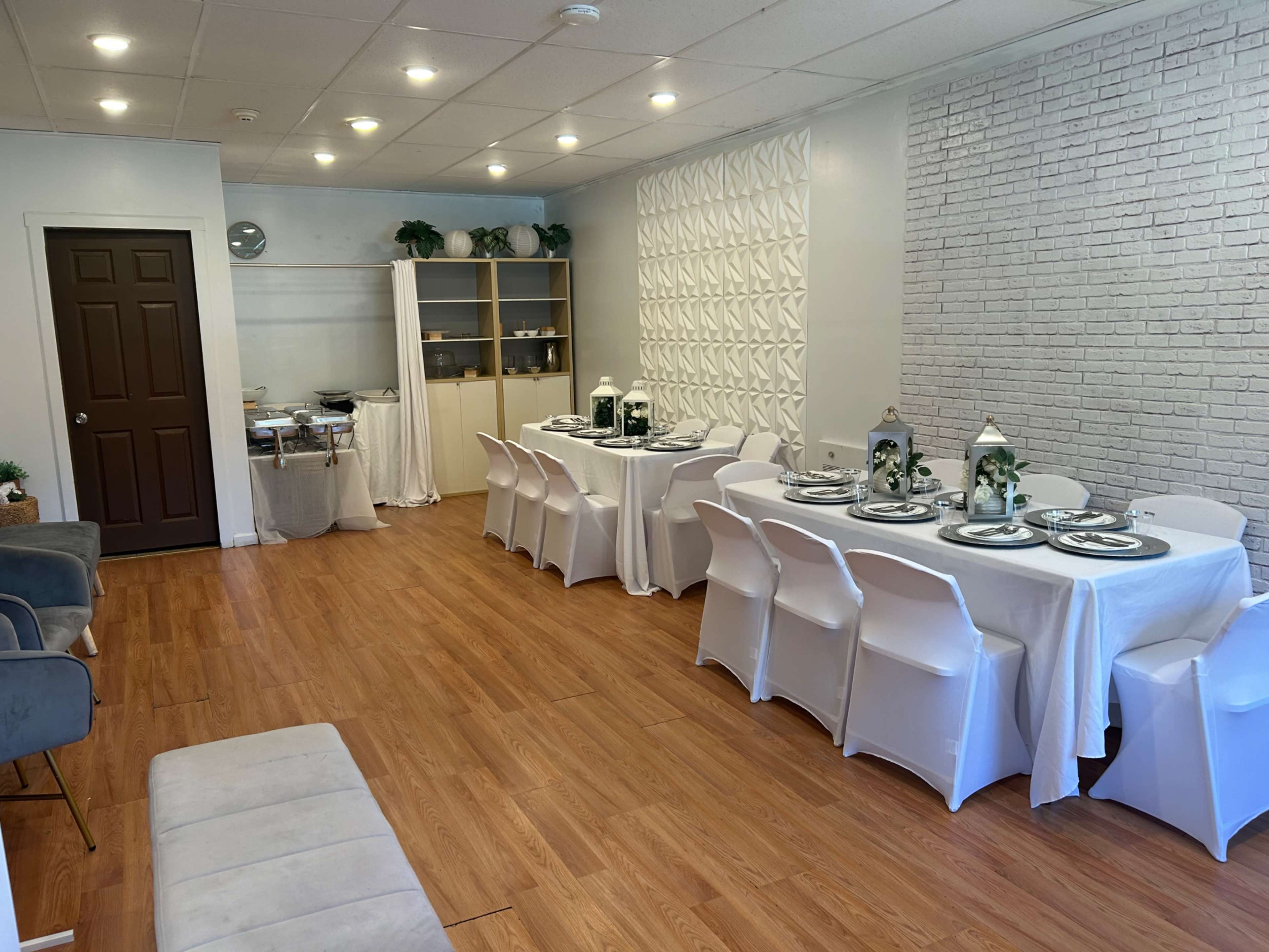 The image shows a bright, modern room furnished with a long dining table set for a meal, surrounded by white chairs, and featuring a buffet setup in one corner.