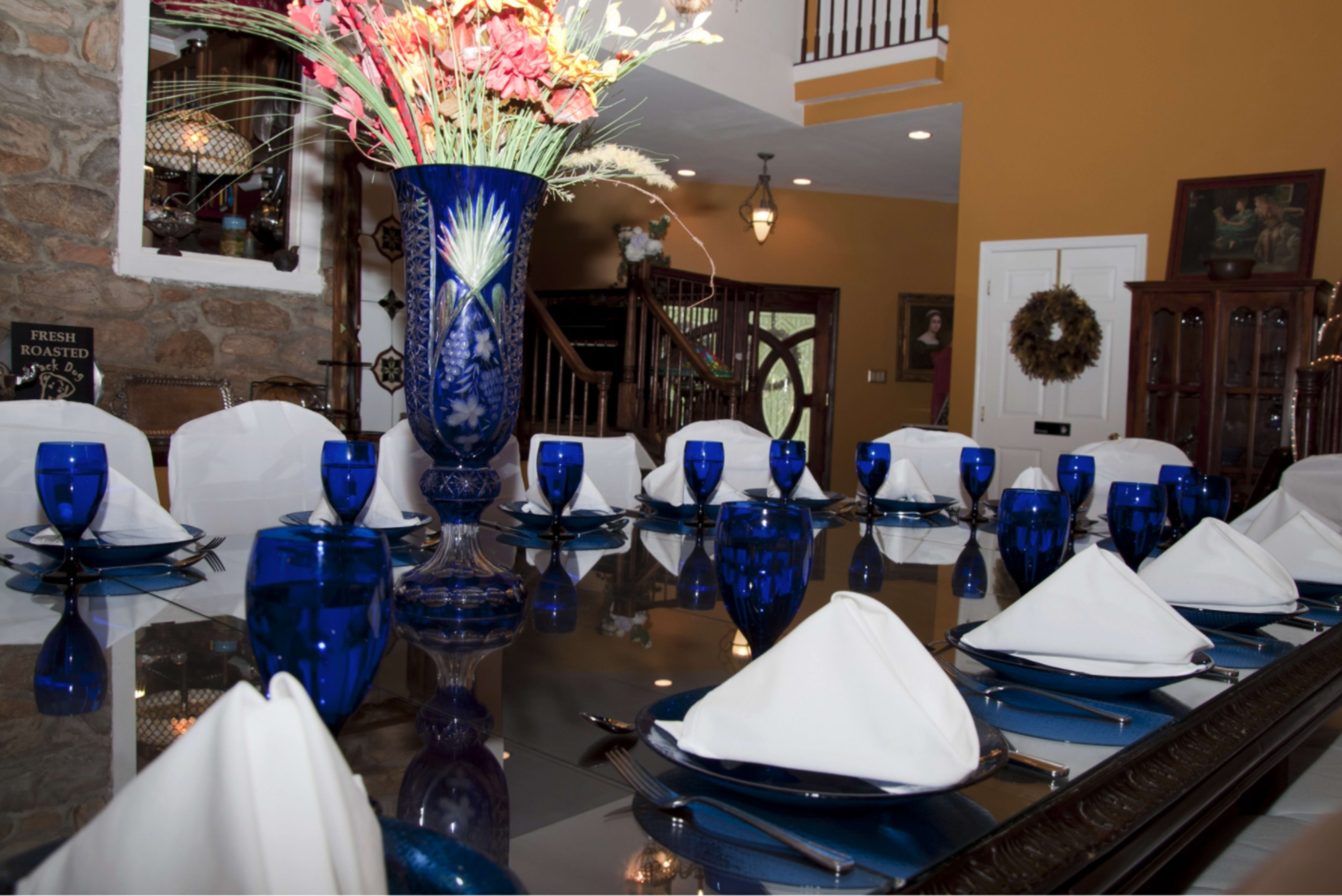 A large dining table is elegantly set with blue glassware and folded white napkins, surrounded by a decorative floral centerpiece and a backdrop of a stone wall and wooden staircase.