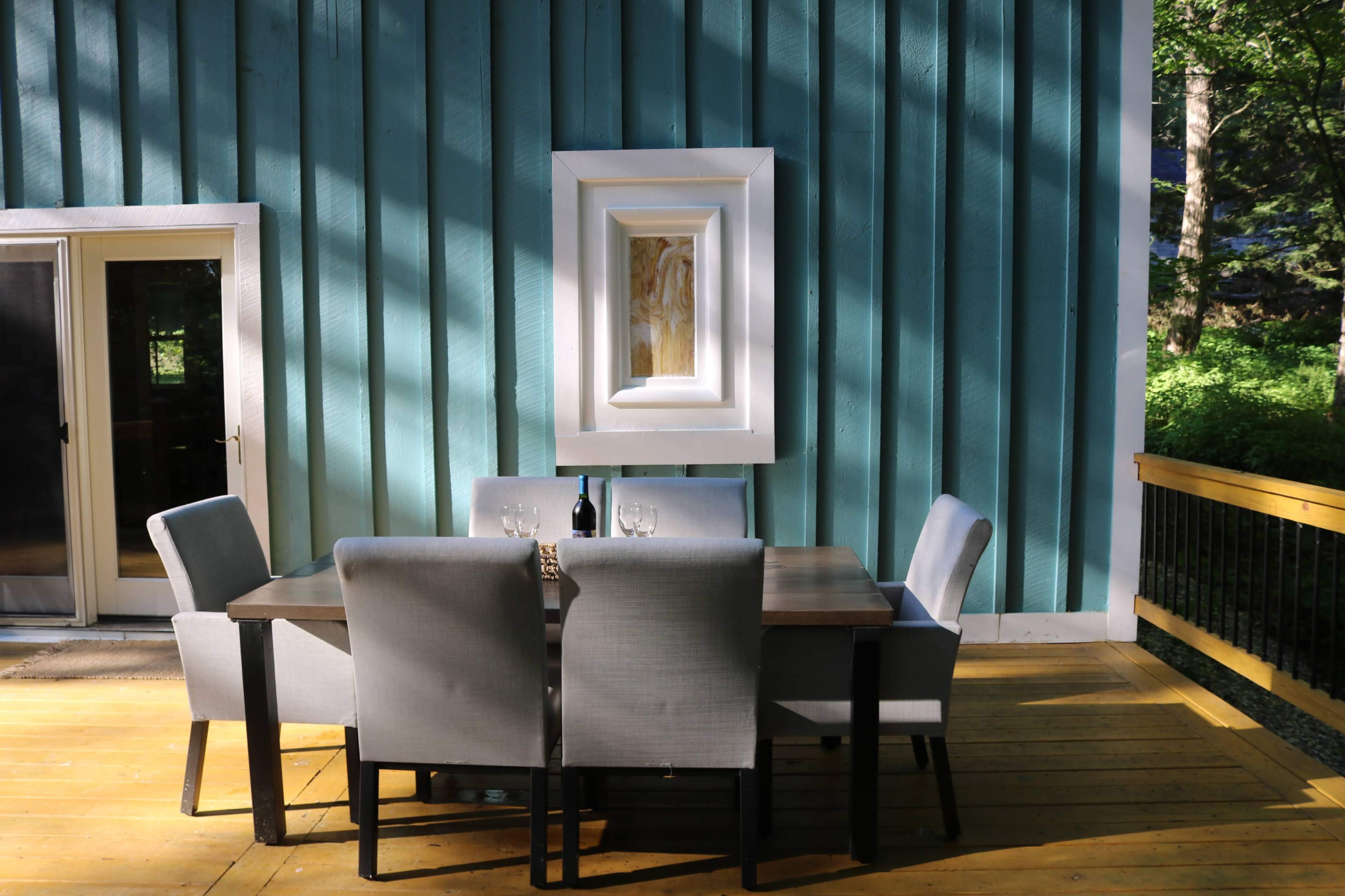 A dining table with gray chairs is situated on a wooden deck against a turquoise wall, featuring a framed painting above the table.