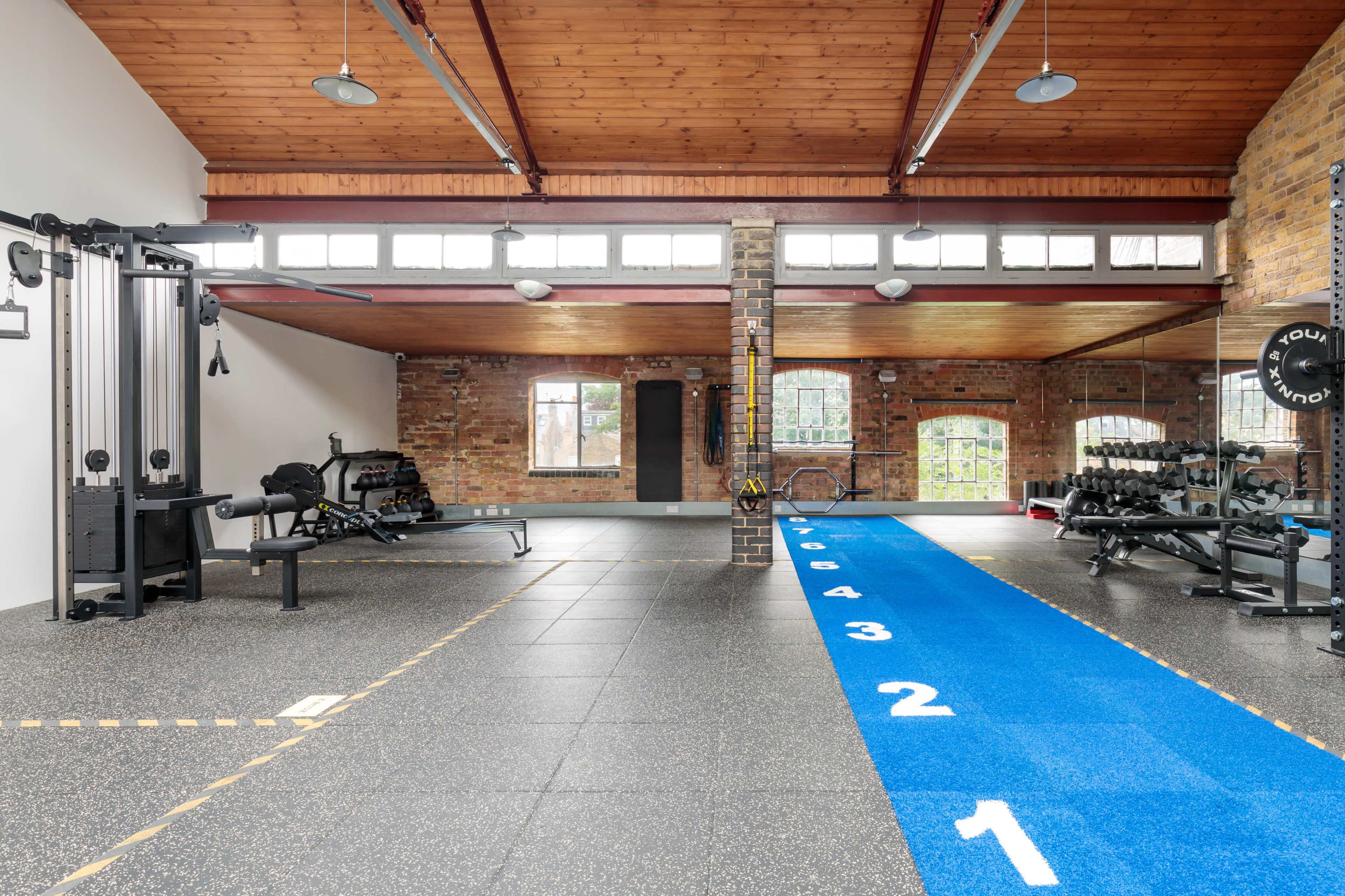 A spacious gym area featuring various workout equipment, a blue track numbered from one to eight, and large windows providing natural light.