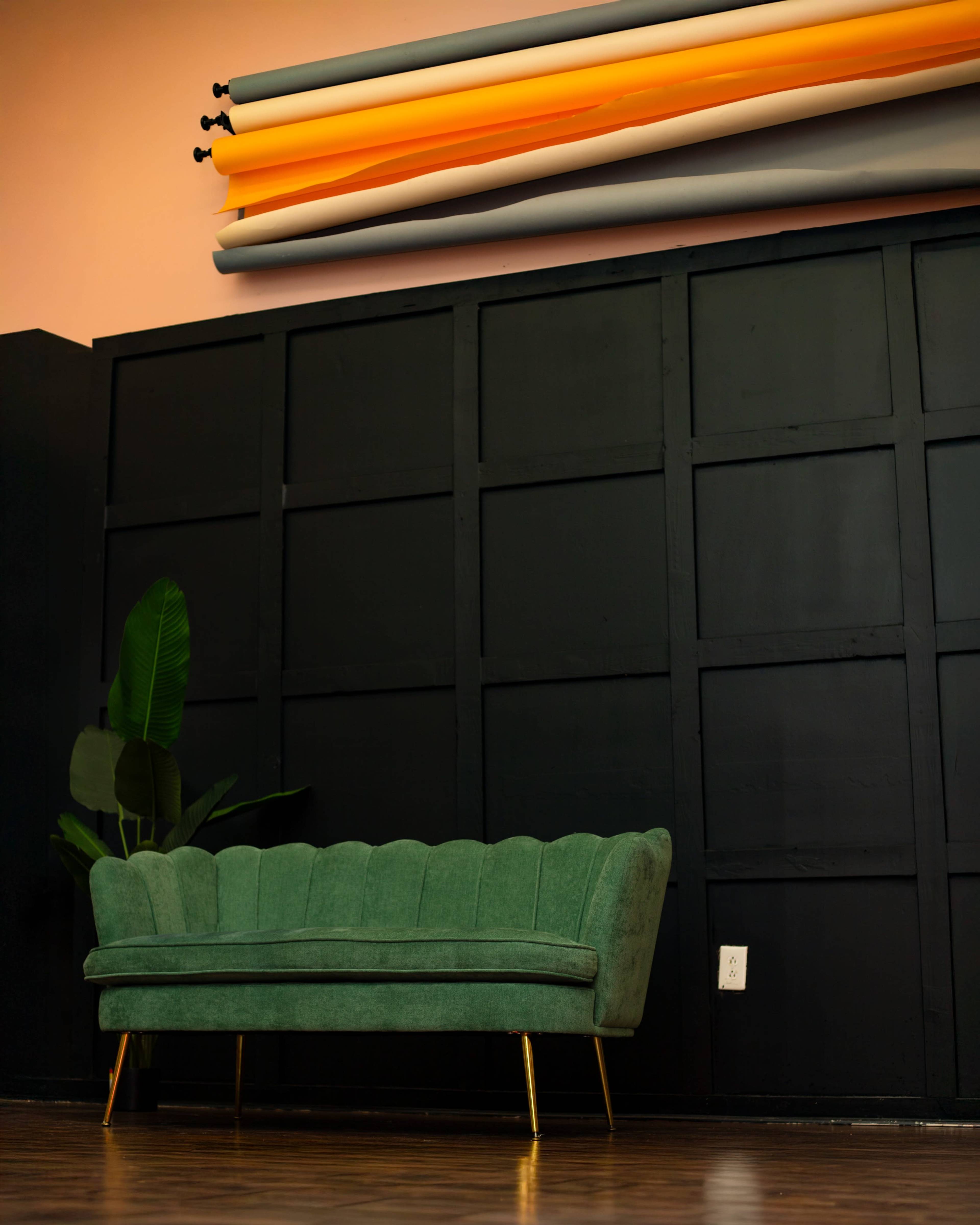The image shows a green velvet sofa with gold legs against a black wall, with rolls of colored paper placed above it.