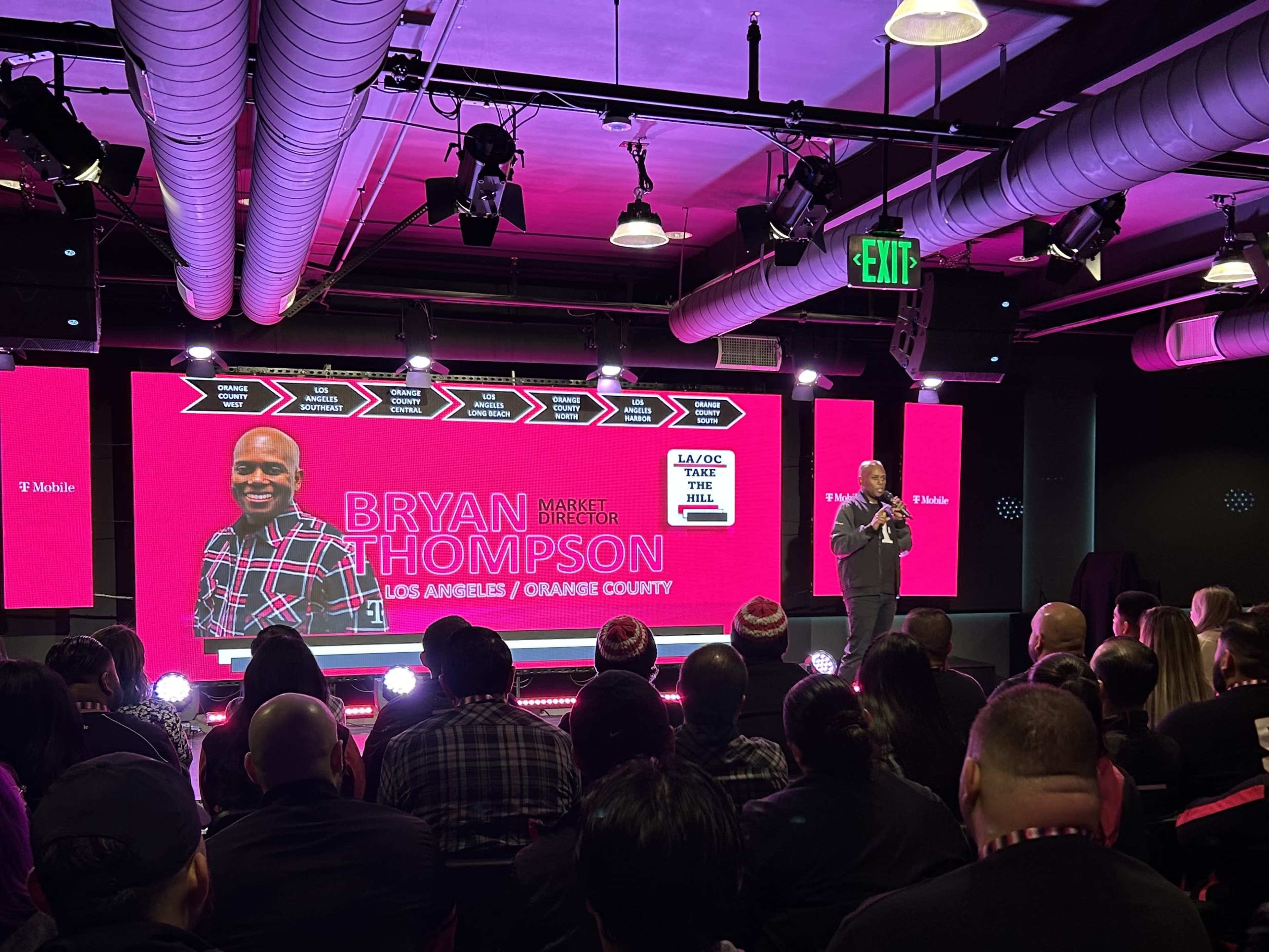 A speaker stands on stage in front of a large screen displaying their name and title during an event with a seated audience.