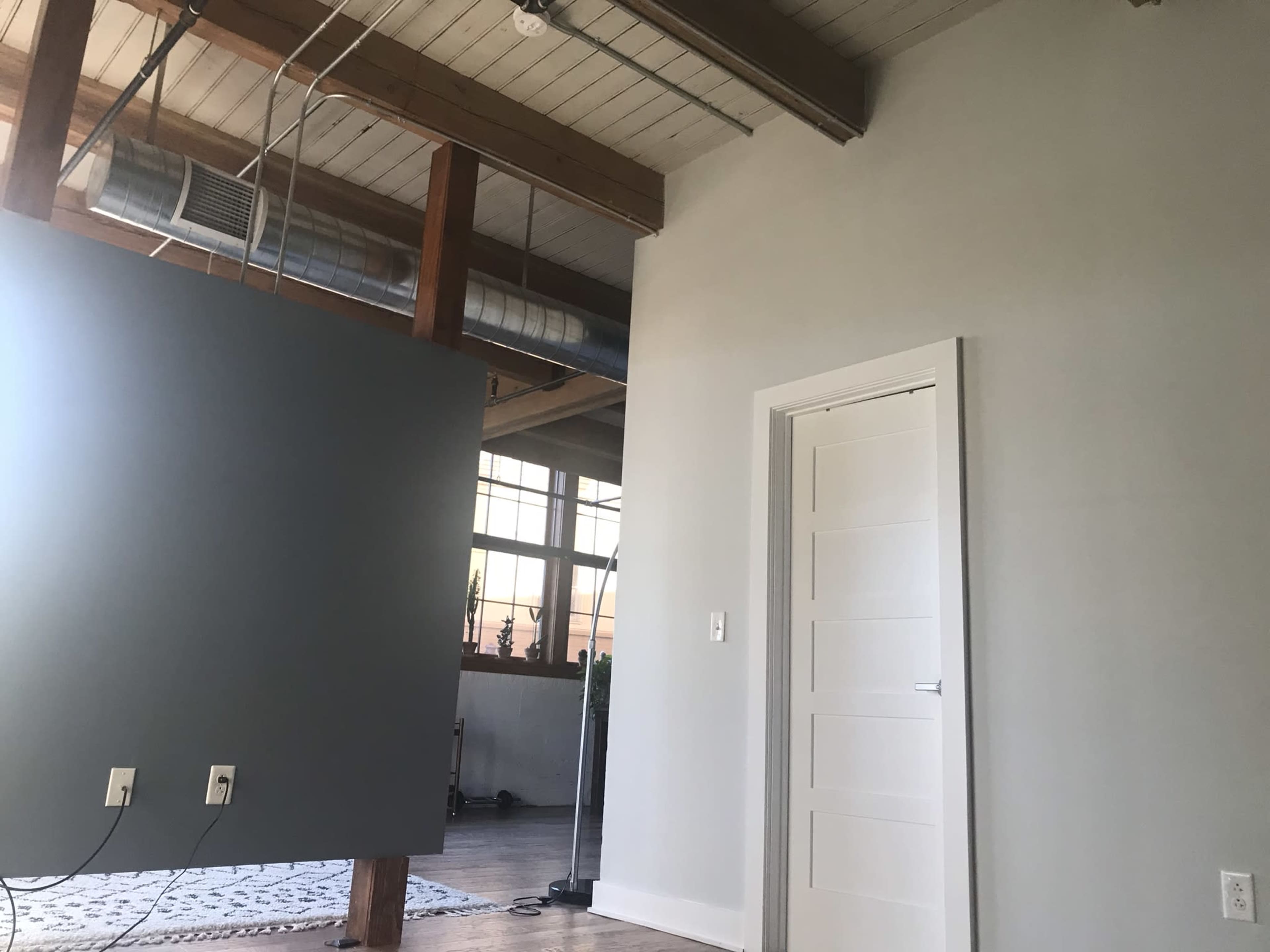 A spacious interior room with a gray wall, a white door, exposed wooden beams, and large windows letting in natural light.