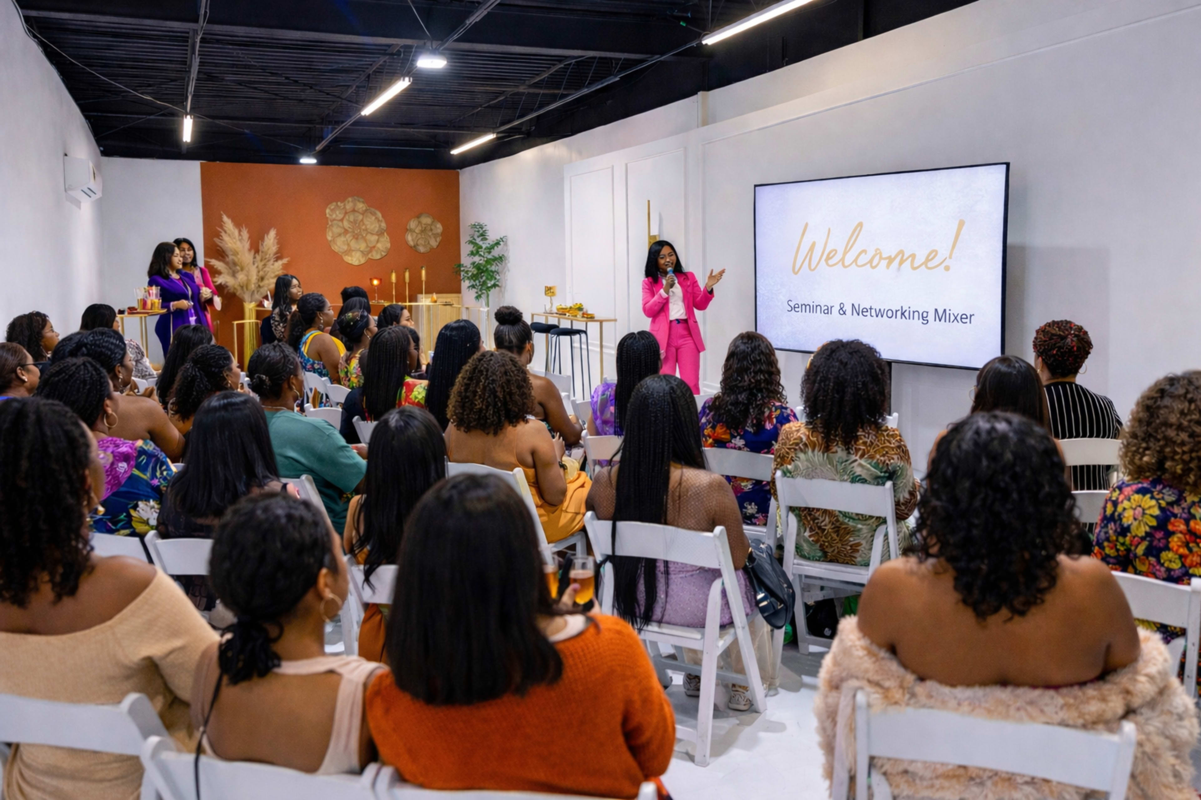 A speaker stands at the front of a seminar room, welcoming a large audience of women seated in white chairs, with decorations and a presentation screen in the background.